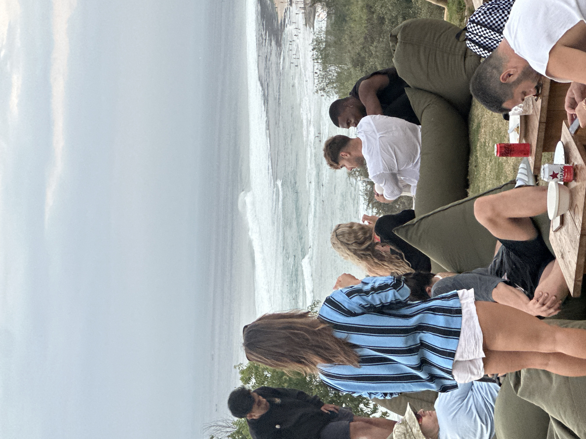 In Kuta Selatan, Indonesia, an outdoor gathering takes place with a group of people relaxing on beanbag chairs, facing a view of the ocean with moderate waves and surfers visible. The sky is overcast with light gray clouds. In the foreground, a woman in a blue and black striped shirt and white shorts stands with her back to the camera. To her left, a man in a black shirt and shorts is partially visible. In the center, two men are seated on large green beanbag chairs; one wears a white shirt and the other a black tank top. Behind them, a woman with blonde hair is seated. To the far right, a man in a white shirt is seated at a wooden table with a checkered bag beside him. On the table, there are plates, bowls, and two cans of drinks. The ground is covered in grass and some fallen leaves.