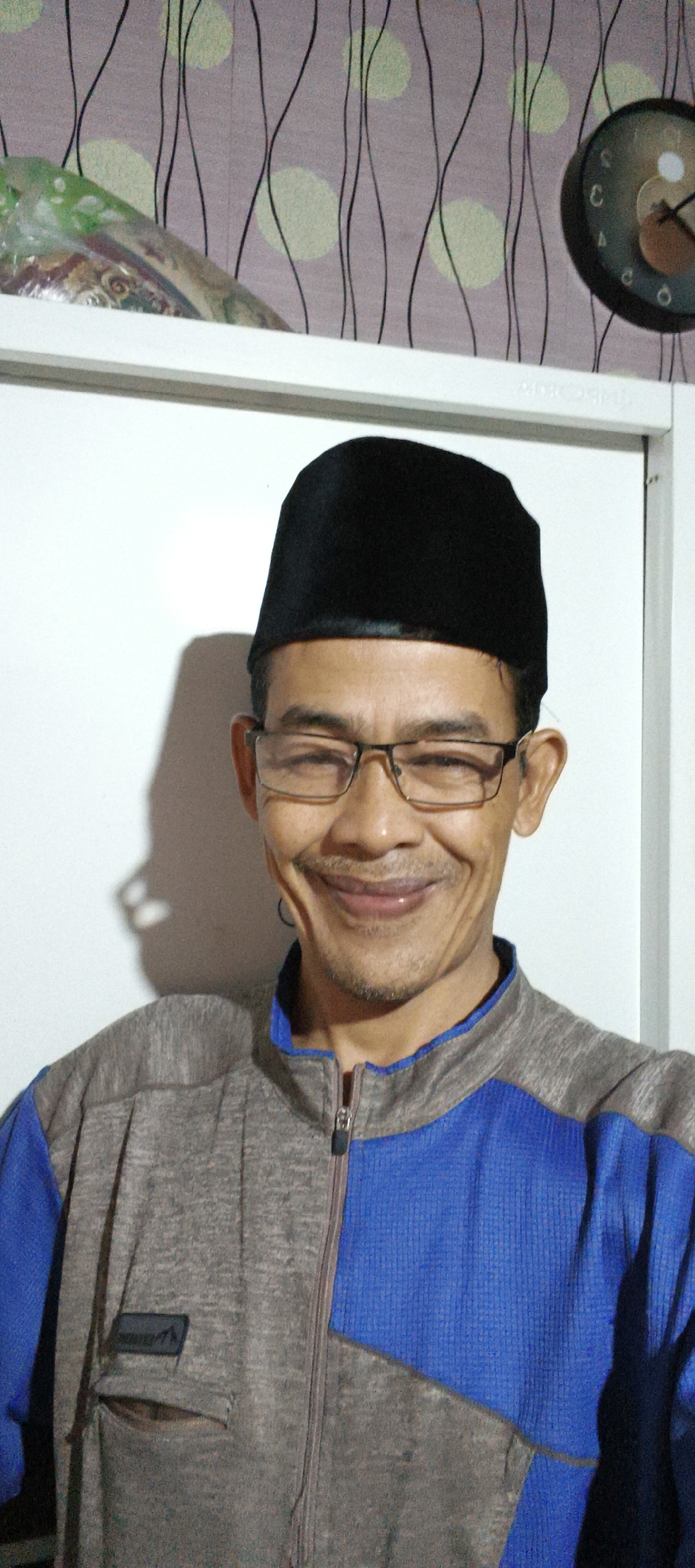 A man wearing a black peci and glasses is looking at the camera, smiling slightly. He is wearing a grey and blue zip-up jacket. The background appears to be a wall with a patterned wallpaper and a clock. The image is taken indoors, likely in a residential setting in Muaro Jambi, Indonesia.