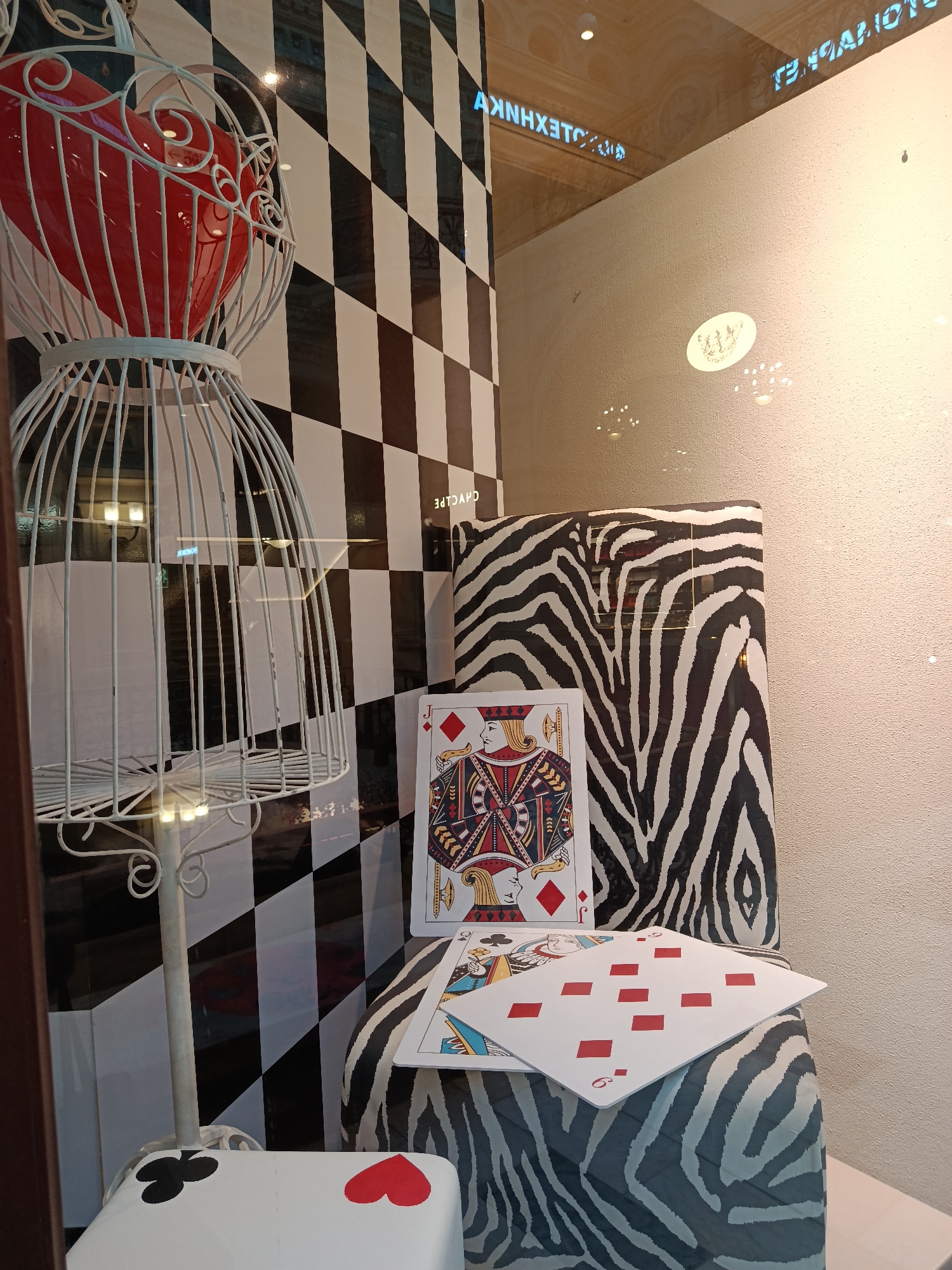 This image captures a vibrant and whimsical shop window display, likely in Moscow, Russia. The scene is dominated by a striking black and white checkered background panel.
In the foreground, a large, ornate white wire structure, resembling a decorative birdcage or a stylized crinoline, encloses a glossy, bright red heart. To the right of this, resting against a black and white zebra-print covered object (possibly a chair or display block), are two oversized playing cards. The most prominent is a King of Diamonds, leaning upright. In front of it, lying flat, is a 6 of Diamonds, though its pips are stylized as rectangular red blocks rather than traditional diamond shapes. Below, in the bottom left corner, a white cube or block is adorned with painted suit symbols: a black club and a red heart.
The display utilizes strong patterns and colors, evoking a sense of fantasy, possibly inspired by "Alice in Wonderland." The time of day appears to be daytime or early evening, with reflections visible on the shop window glass. These reflections include reversed Cyrillic text, such as "ТЕХНОЛОГИЯ" (Technology) and partial words like "AKNHXETC" and "CHAVCHE," suggesting storefronts or signs from across the street or behind the viewer. A small, indistinct circular logo or emblem is also reflected on the upper right portion of the glass. The overall impression is one of an imaginative and eye-catching retail presentation.
