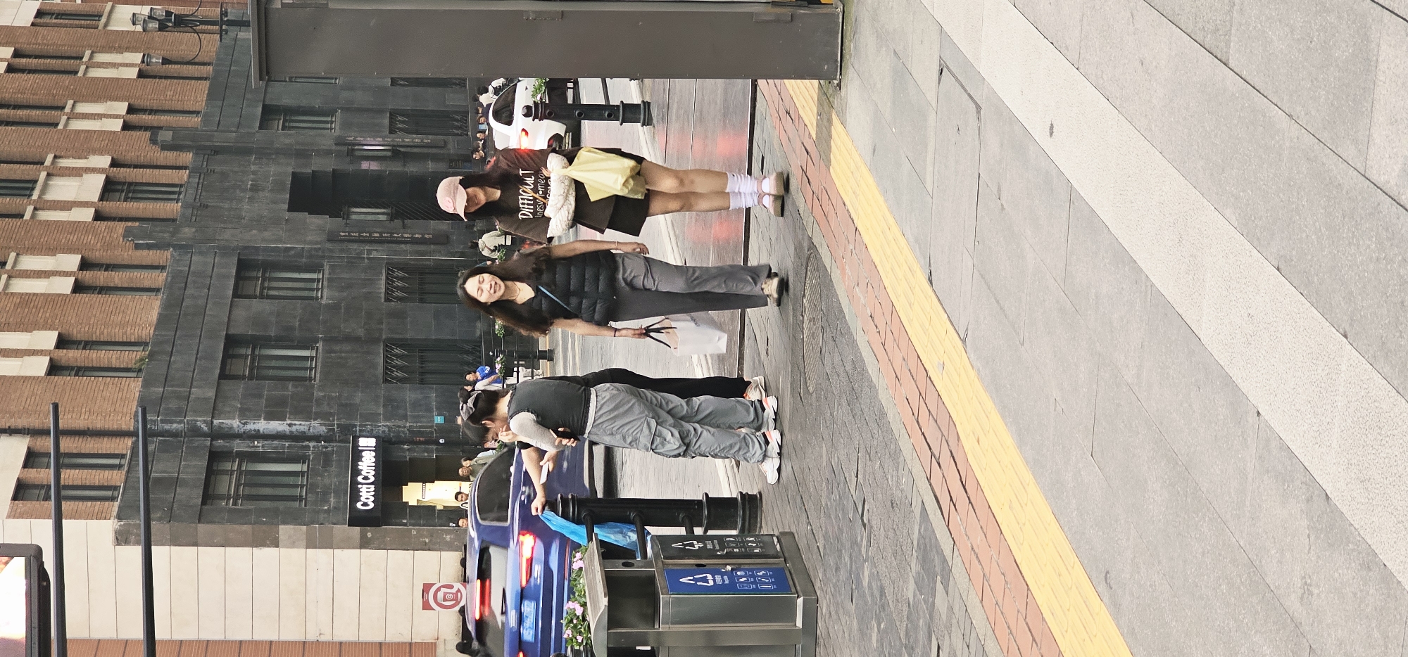 A street scene in Yuzhong District, China, shows pedestrians on a sidewalk. A building with "Cotti Coffee" signage is visible. Two young women are standing together while an individual is putting trash in a nearby trash receptacle. Cars are passing by. The sidewalk has a colored safety strip. The image is taken at an angle, with the street receding into the distance.