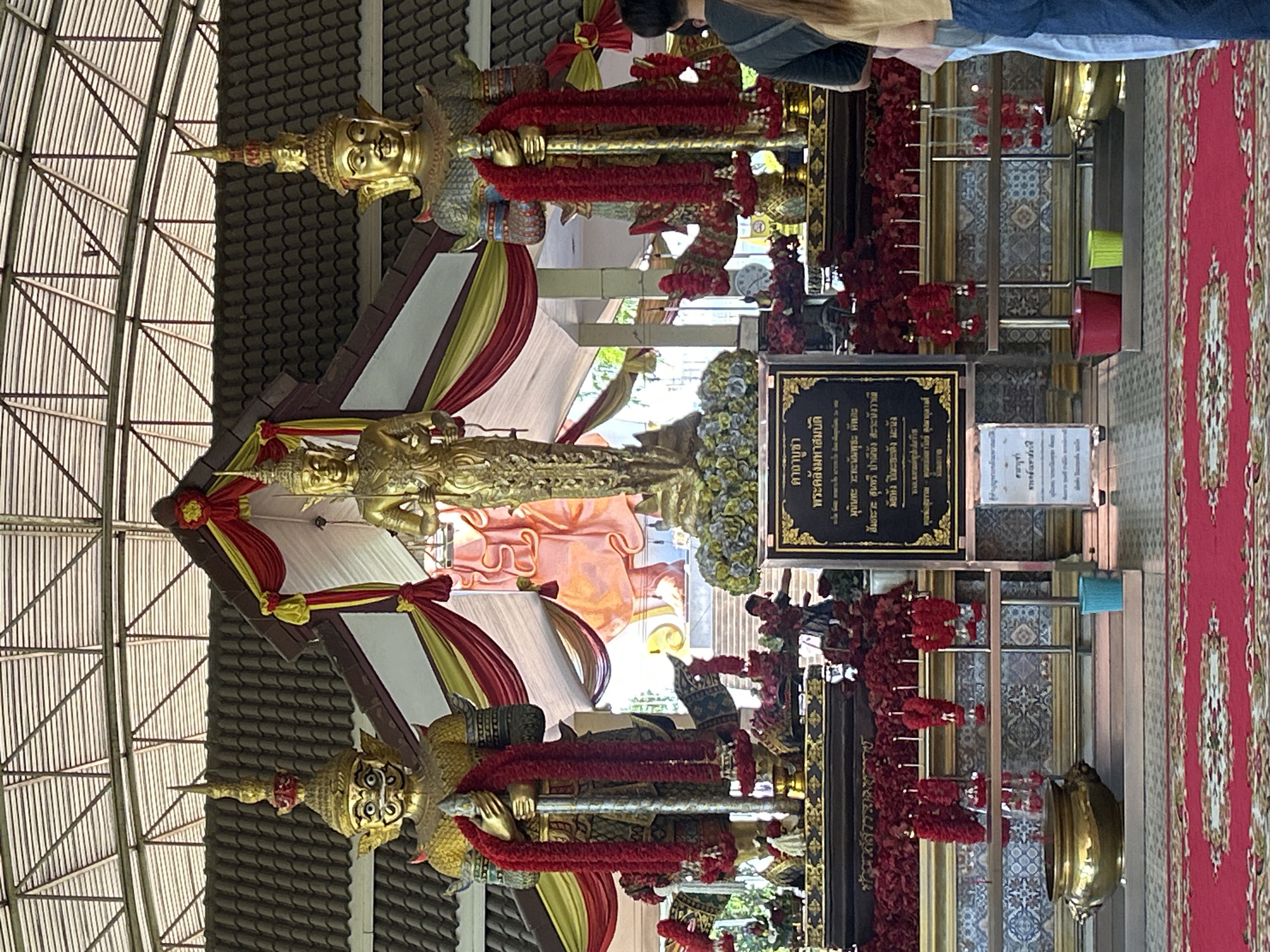 The image shows a shrine with three golden statues. Two large warrior-like statues flank a central, slender statue. Both large statues are adorned with red garlands. A black sign with Thai script is placed in front of the central statue. The shrine is decorated with red and yellow draped fabric. In the background, a large, pale pink statue is visible. Red flowers are arranged in front of the statues. Several golden bowls and buckets are present in the foreground and on tiered platforms. The scene is covered by a roof structure supported by beams. Two individuals are partially visible on the right side of the image, one facing away from the camera and the other facing towards the shrine. The ground is covered by a red carpet with a patterned design. Based on the Thai script on the sign, the shrine is located at Wat Phra That Khao Noi in Thai Ko Subdistrict, Mueang Nan District, Nan Province, Thailand.