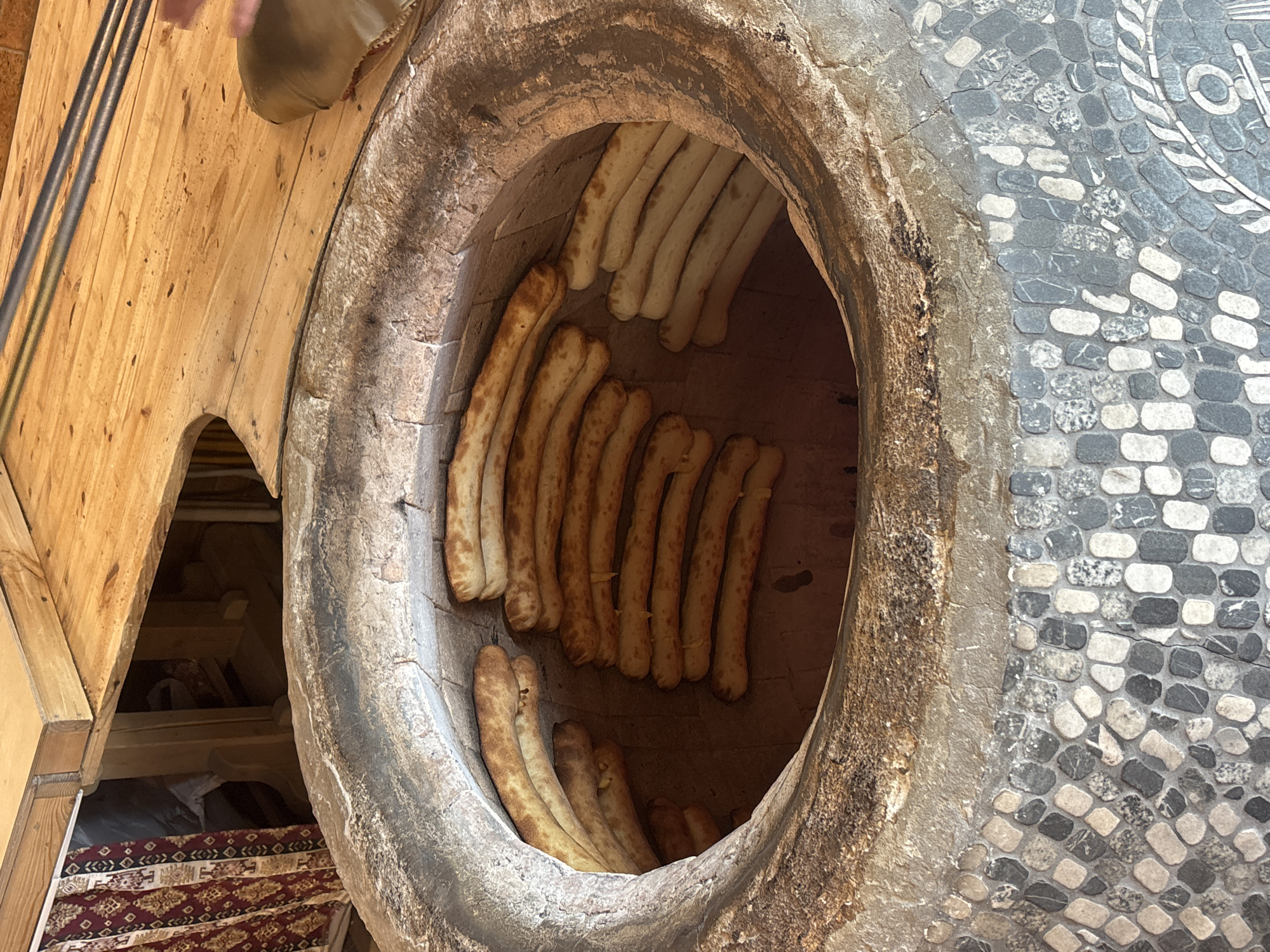 The image shows a tandoor oven with Armenian bread, likely in Tsovagyugh, Armenia. Long loaves are baked vertically against the interior walls of the oven. The exterior of the oven appears to be constructed of stone and is located indoors. Adjacent to the oven is wooden structure. A person's foot is visible at the top left. To the right is a mosaic stone pattern. A rug is visible below and to the left of the wooden structure.