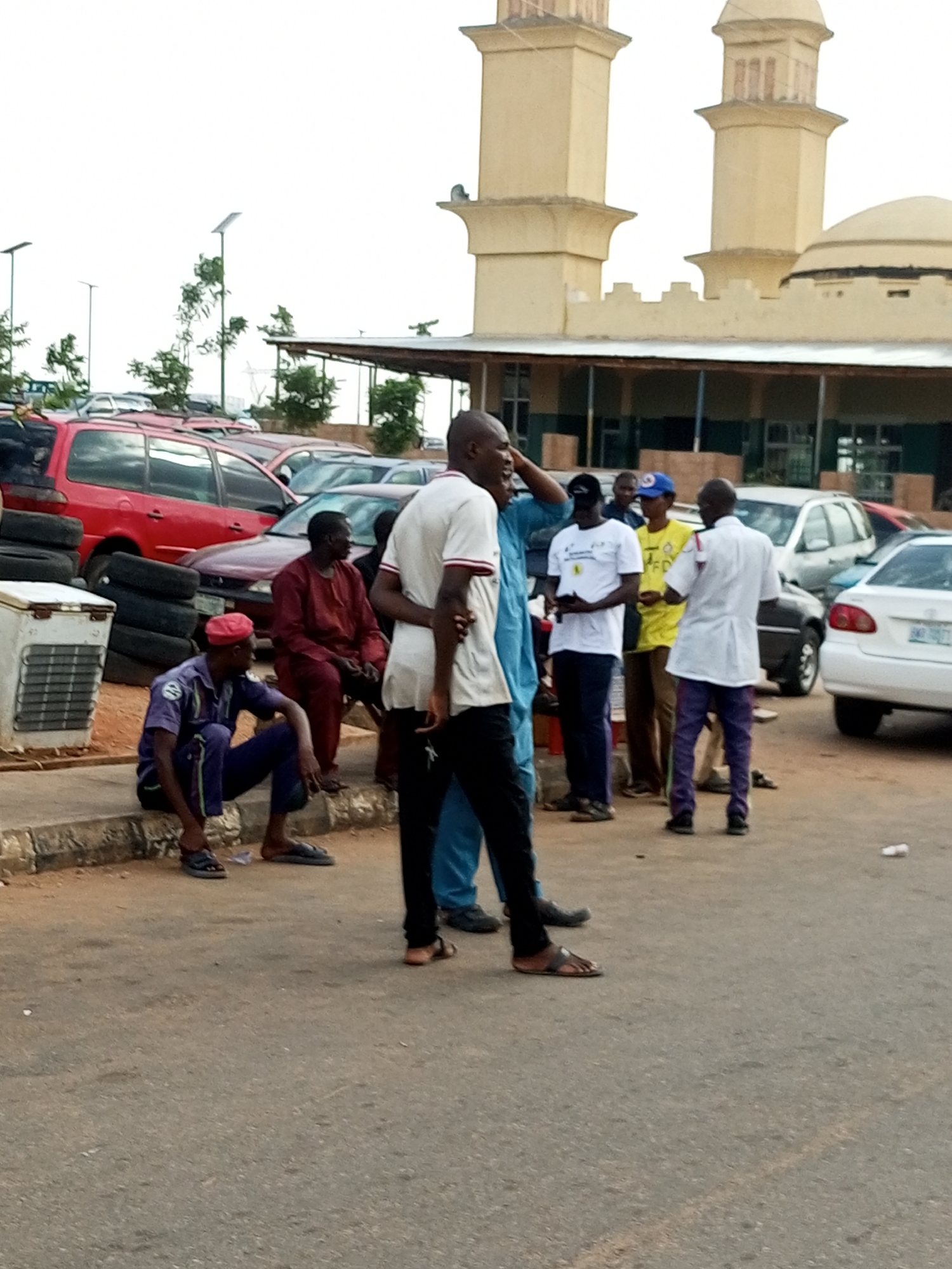 The image depicts a group of men gathered outdoors, with a mosque-like building visible in the background. In the foreground, a man in a white shirt and black pants is engaged in conversation with another man. To the left, a man in a purple uniform with a red cap is seated on a curb. Behind him, a man in a maroon robe is also seated. Several vehicles, including a red SUV and other cars, are parked in the vicinity. Stacks of tires and an air conditioning unit are also present. The environment appears to be a street or parking area, with a paved surface and a concrete curb. The sky is overcast.