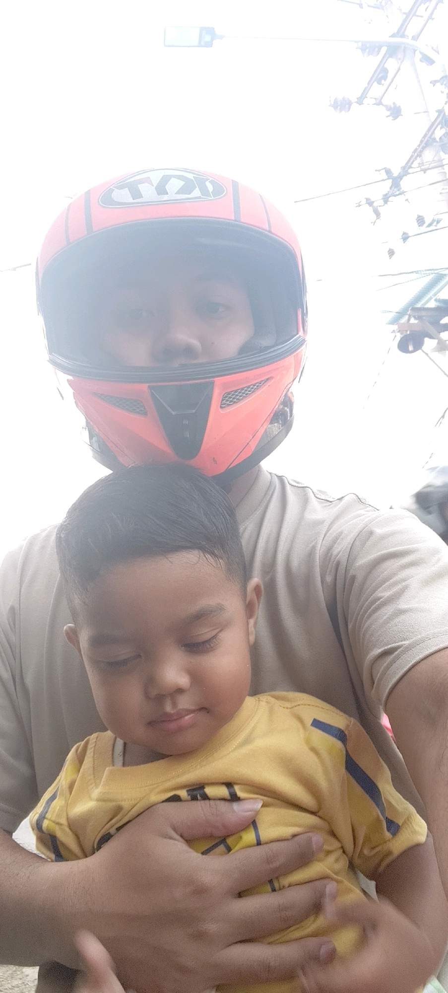 A close-up, vertical, eye-level selfie taken from a motorcycle shows two individuals. The person in the foreground, presumably the driver, is wearing a bright orange motorcycle helmet with black accents and the "KYT" logo visible. The helmet obscures most of their face, but their eyes and the bridge of their nose are visible. They are wearing a light brown t-shirt.
Held by the driver in the foreground is a young child, likely a boy, wearing a yellow t-shirt with dark blue stripes on the sleeves. The child's head is tilted downwards, with eyes closed and a slight smile on their lips. Their dark hair is neatly styled. The child is being cradled by the driver's arms and hands, which are also visible in the lower portion of the image. The background is blurred and overexposed, showing a bright sky with what appears to be utility poles and wires, suggesting an outdoor setting in an urban or semi-urban environment. The scene is consistent with a photo taken while riding a motorcycle in the general vicinity of Mekarmukti, Indonesia, which has similar infrastructure and common transportation modes.