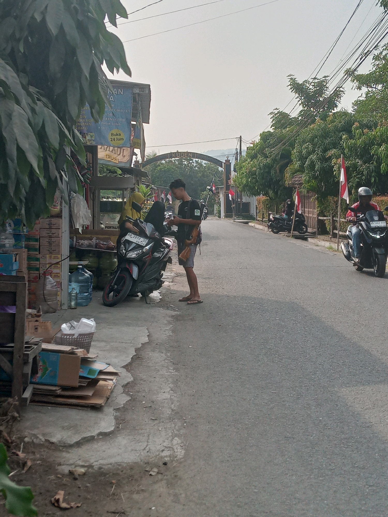 The image depicts a street scene in Sitimulyo, Indonesia, likely a commercial area. In the foreground, a young man in a dark t-shirt and shorts stands beside a black motorcycle, looking at his phone. To his left, a woman in a yellow headscarf and dark clothing is seen near the same motorcycle, which is parked on the side of a paved road. Various items, including boxes, water gallons, and a basket of what appears to be plastic bags, are stacked on the sidewalk next to a small building with signage. The building has a blue sign with white text, possibly indicating services offered.
Further down the road, another person is riding a black motorcycle, wearing a helmet and a red jacket. A third black motorcycle is parked further in the distance. Indonesian flags are visible, some mounted on poles and others attached to the fence. Lush green trees line the right side of the road, and an archway with decorative elements is visible in the background, possibly marking an entrance to a village or area. The sky is overcast, suggesting a hazy or cloudy day. The overall environment appears to be a typical street in a tropical region, with a mix of residential and commercial elements.