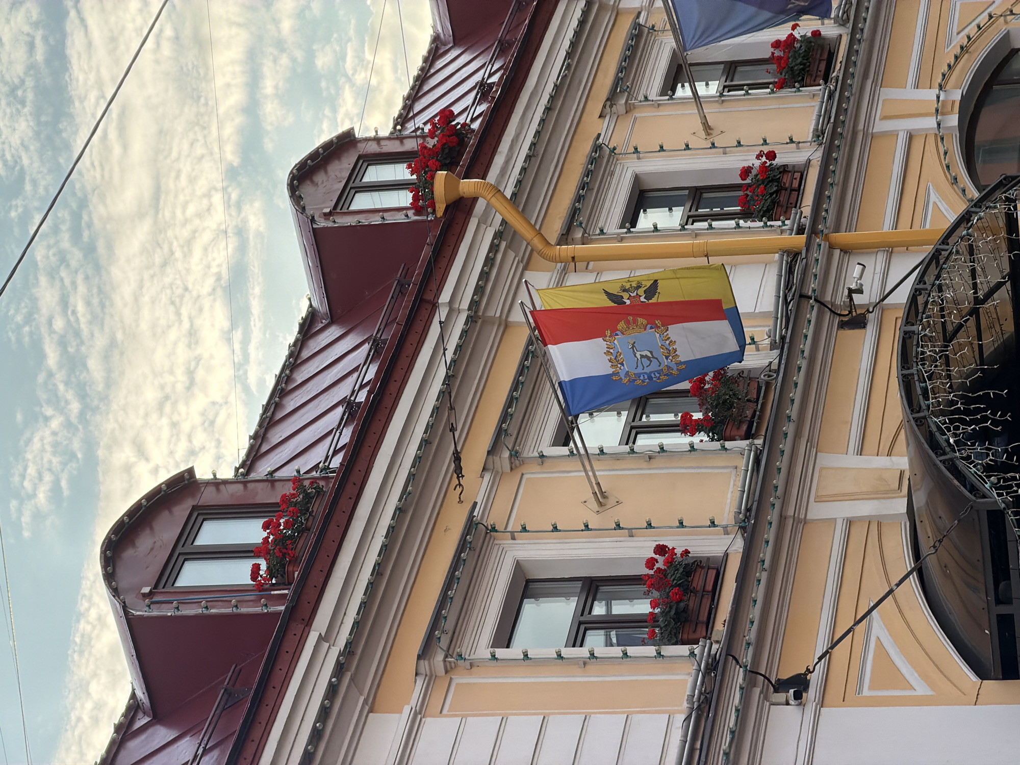 A building facade in Saint Petersburg, Russia, features a multi-story structure with a reddish-brown mansard roof adorned with decorative lights. The building has a pale yellow painted facade with white trim around the windows. Several windows are visible, some with flower boxes containing red flowers. A yellow drainpipe runs down the side of the building. Three flags are displayed horizontally, with the leftmost flag being tricolor with a crest. The sky above is partly cloudy with a pale, diffused light. The image captures a street-level view looking upwards at the building.