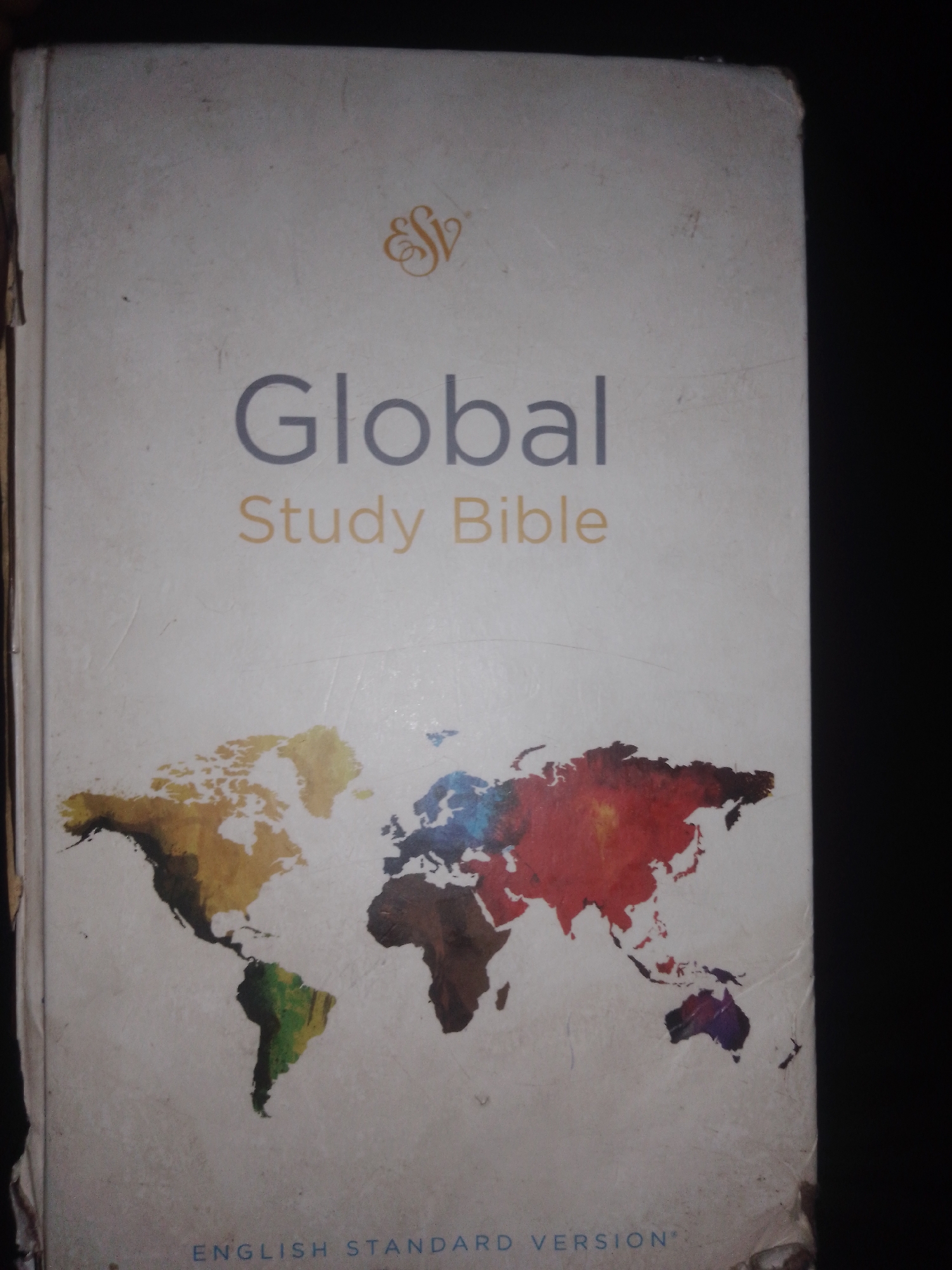 The image is a close-up, high-angle, outdoor shot of the cover of a book titled "Global Study Bible" in English Standard Version. The book cover is a light beige color and features a stylized logo at the top, followed by the title in gray and yellow text, and a colorful, abstract world map taking up the majority of the lower half of the cover. The book is standing upright, leaning slightly to the left, against a dark background that suggests it is placed on a surface or against a wall. The book cover shows some signs of wear, with scuff marks and minor damage visible along the edges. The lighting is dim, casting a shadow on the right side of the image. There are no people or animals visible in the image. The location of Jos, Nigeria is not evident from the image itself; this context is external information.