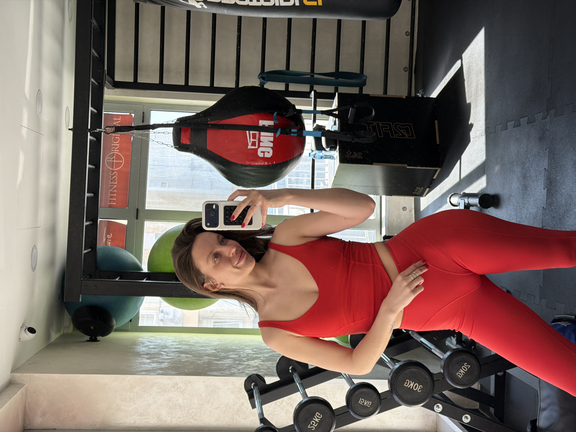 The image displays an adult female taking a mirror selfie inside the "Fitness Original" gym, located in Budva, Montenegro. The subject, with long brown hair, wears a red athletic crop top and matching leggings. She holds a white smartphone with a light blue case in her right hand, positioned to capture her reflection. Her left hand rests near her hip, adorned with a silver-colored ring. Her fingernails are painted red, and she wears small earrings, exhibiting a subtle smile.
The gym environment features black interlocking rubber floor mats and light-toned, textured walls. A white dome security camera is mounted on the ceiling in the upper left. Through large windows in the background, a reversed red "Fitness Original" sign is visible, confirming the gym's identity and location, with exterior buildings discernible beyond.
Visible gym equipment includes: a black metal dumbbell rack on the left with circular weights, including denominations such as "30KG" and "12KG" (reversed in the mirror); two large stability balls (one teal, one green) stored on a rack; a black and red tear-drop shaped punching bag, marked "KING" (reversed), suspended from a black metal frame; a black cylindrical punching bag with white lettering to the right; a black wooden plyometric box beneath the tear-drop bag, holding a plastic water bottle; blue and black TRX suspension straps; and a blue resistance band hanging on the wall or frame. Sunlight casts distinct shadows from the window frames and equipment onto the floor.
