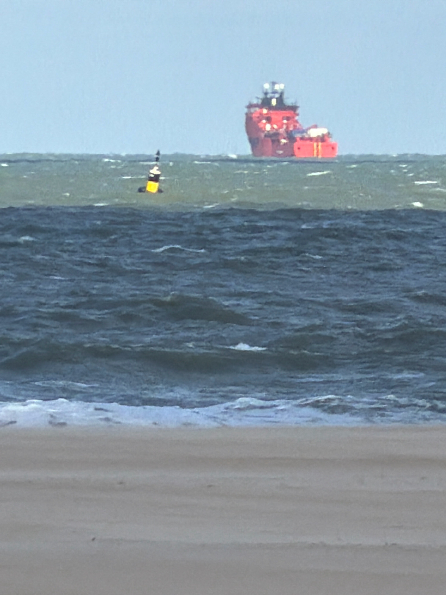 An extreme long shot shows a choppy sea under a clear sky with a red ship on the horizon and a yellow and black buoy in the mid-ground. The foreground shows a sandy beach with a few faint footprints. The scene is likely the coast of Ostend, Belgium, given the presence of a buoy, common in port areas.