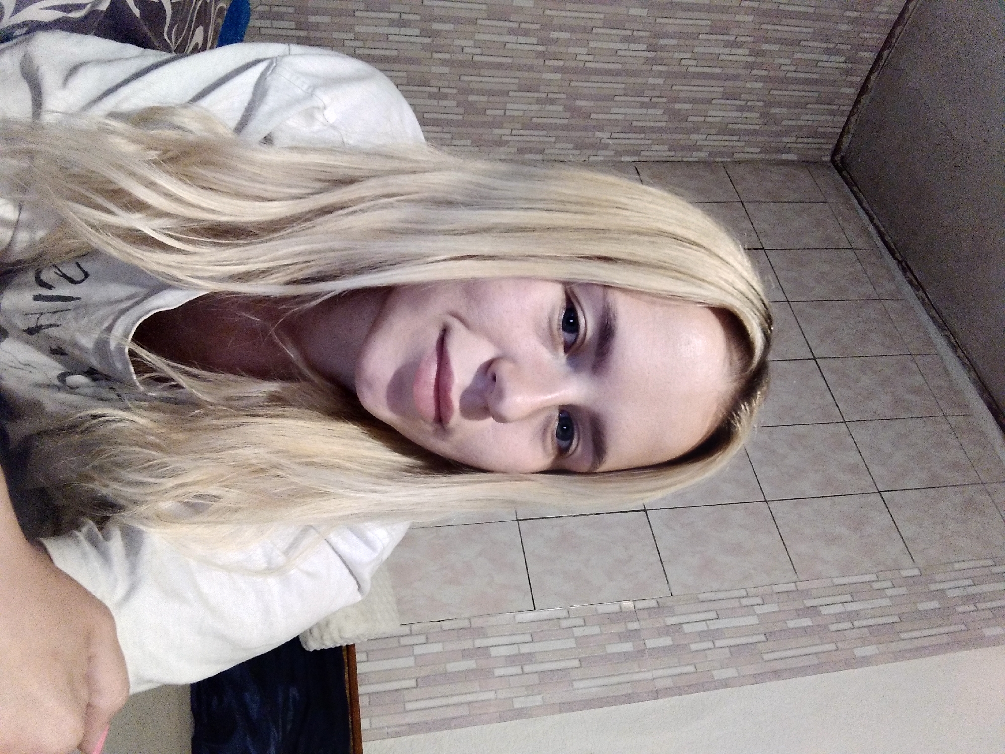 A young woman with blonde hair pulled back in a ponytail is looking at the camera. She is wearing a white t-shirt. The background appears to be a bedroom with tiled walls. The lighting is dim and the overall atmosphere is casual.