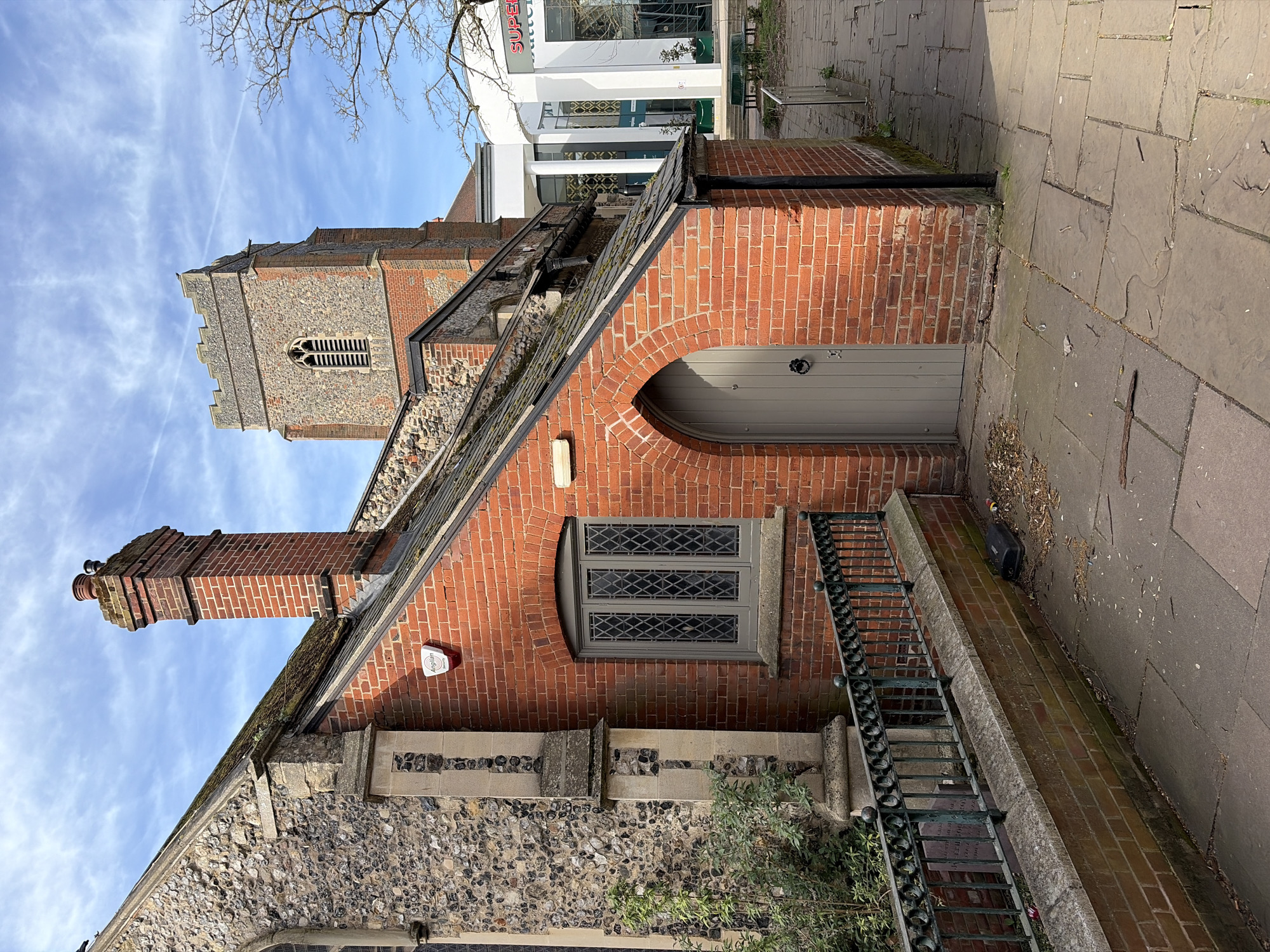 This image captures a section of St. Margaret's Church in Ipswich, United Kingdom. The scene features a brick-built outbuilding connected to the main church structure. The outbuilding has a distinctive arched doorway and a leaded window. A brick chimney rises from the outbuilding's roof. The church tower is visible in the background against a partly cloudy sky. A paved walkway runs adjacent to the building, with metal railings separating it. The overall scene is one of historical architecture in an urban setting.
