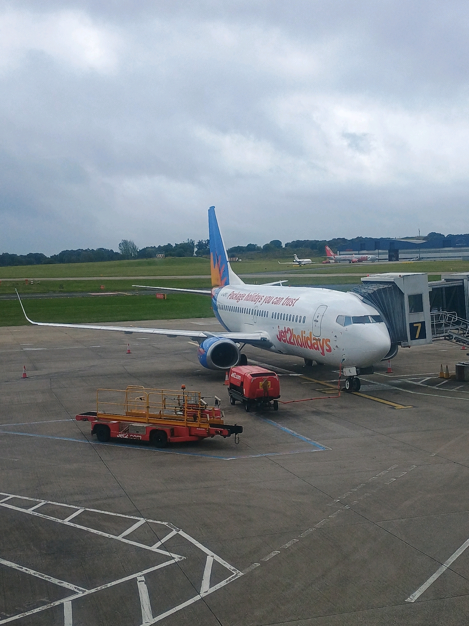 The image depicts an airport scene on an overcast day. A Jet2.com Boeing 737 aircraft is parked at a gate, connected to a jet bridge labeled with the number '7'. The plane is white with "Jet2holidays" branding and a colorful tail fin. In the foreground, a red baggage tug with yellow accents and "Jet2.com" written on it is positioned on the tarmac. Other airport equipment and markings are visible on the concrete surface. In the background, several other aircraft can be seen on the runway and taxiway, along with airport buildings and trees. The overall environment is that of an operational airport.