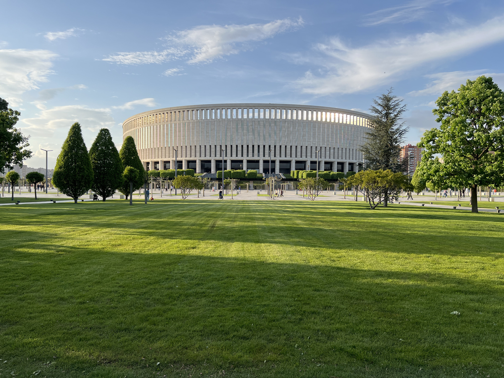 The image shows Krasnodar Stadium, also known as the Colosseum. It's a sunny day with a few wispy clouds in the sky. A large expanse of bright green grass lawn extends toward the circular stadium. The grass is neatly mowed with light and shadowed strips. The stadium itself is a cylindrical structure, surrounded by a colonnade of closely spaced columns. Between the columns are large glass windows for the executive seating and suites. The lawn is surrounded by various trees and shrubs, including conical evergreen trees and rounded deciduous trees. People are walking around the perimeter of the stadium at a distance. The image is taken at ground level.