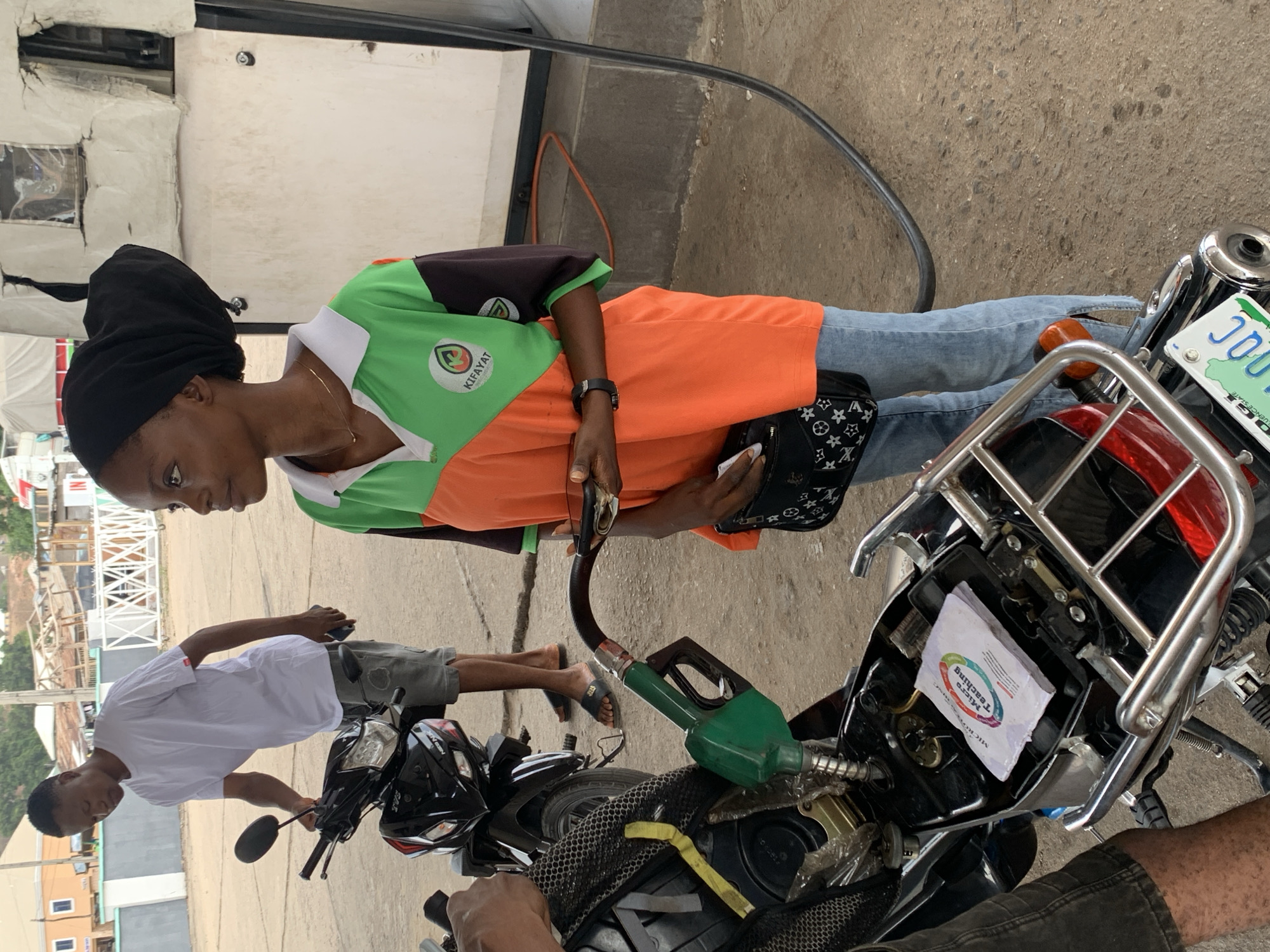 Here is a detailed description of the image:
**Setting:** The scene appears to be a gas station in Lokoja, Nigeria. The image shows a gas pump, concrete pavement, and a building in the background with white siding and windows. There is a view of an outdoor environment with structures and vegetation in the background.
**People:**
* One woman is visible, wearing a black head covering, a shirt that is green with a logo on it, orange, and black in color, and jeans. She is holding a gas pump nozzle and a small patterned purse.
* There is also a man in a white shirt and grey shorts standing near a motorcycle, possibly attending to it or waiting.
**Objects:**
* Two motorcycles are present. One is being filled with gasoline.
* A gas pump with a green nozzle and hose.
* A purse held by the woman.
* A license plate on one of the motorcycles, showing "JOU" and "Nigeria."
* A white paper product in the storage area of the motorcycle being refueled.
**Actions:**
* The woman is actively pumping gasoline into a motorcycle.
* The man is standing nearby the motorcycles.
**Interactions:**
* The woman is engaged in the action of fueling the motorcycle.
* The man nearby is possibly associated with the motorcycle, perhaps waiting for it to be filled up.
**Environment:**
* The scene is well-lit, suggesting it is daytime.
* The presence of the gas pump and vehicles indicates a commercial or service-oriented location.
* The environment appears to be in a typical urban setting.