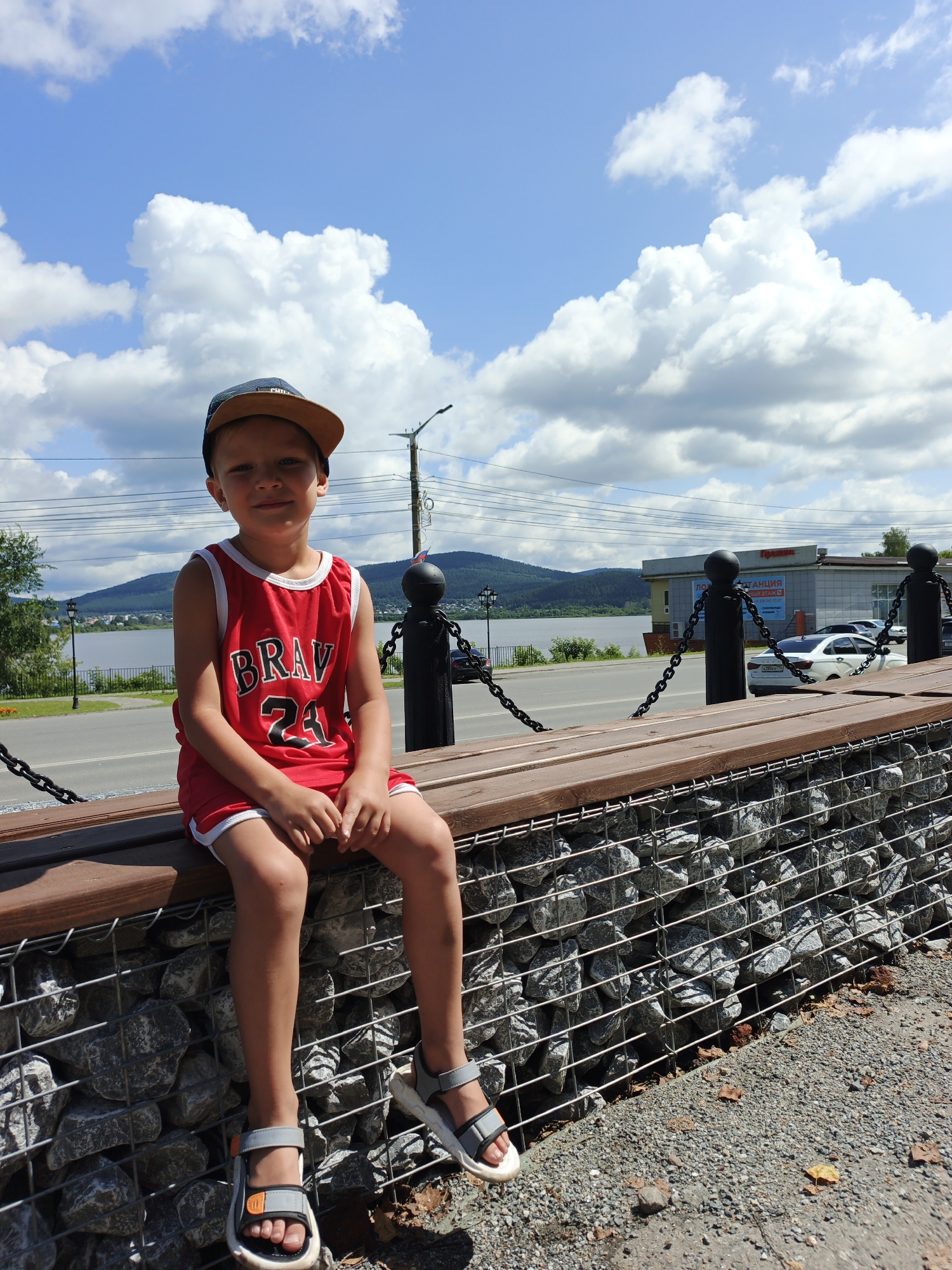 A young boy is sitting on a wooden bench with a gabion wall base, looking towards the camera with a slight smile. He is wearing a red basketball jersey with "BRAV 24" printed in white, a baseball cap, and grey sandals. The bench is situated along a waterfront promenade, with a road and buildings in the background. The sky is blue with fluffy white clouds, suggesting a sunny day. The boy appears relaxed and happy. In the distance, a body of water is visible, with hills and trees on the opposite shore. The scene captures a casual, outdoor moment, likely during daylight hours given the bright sunlight and clear sky. Text on a building in the background appears to be in Russian. The presence of a road and buildings suggests an urban or semi-urban environment.