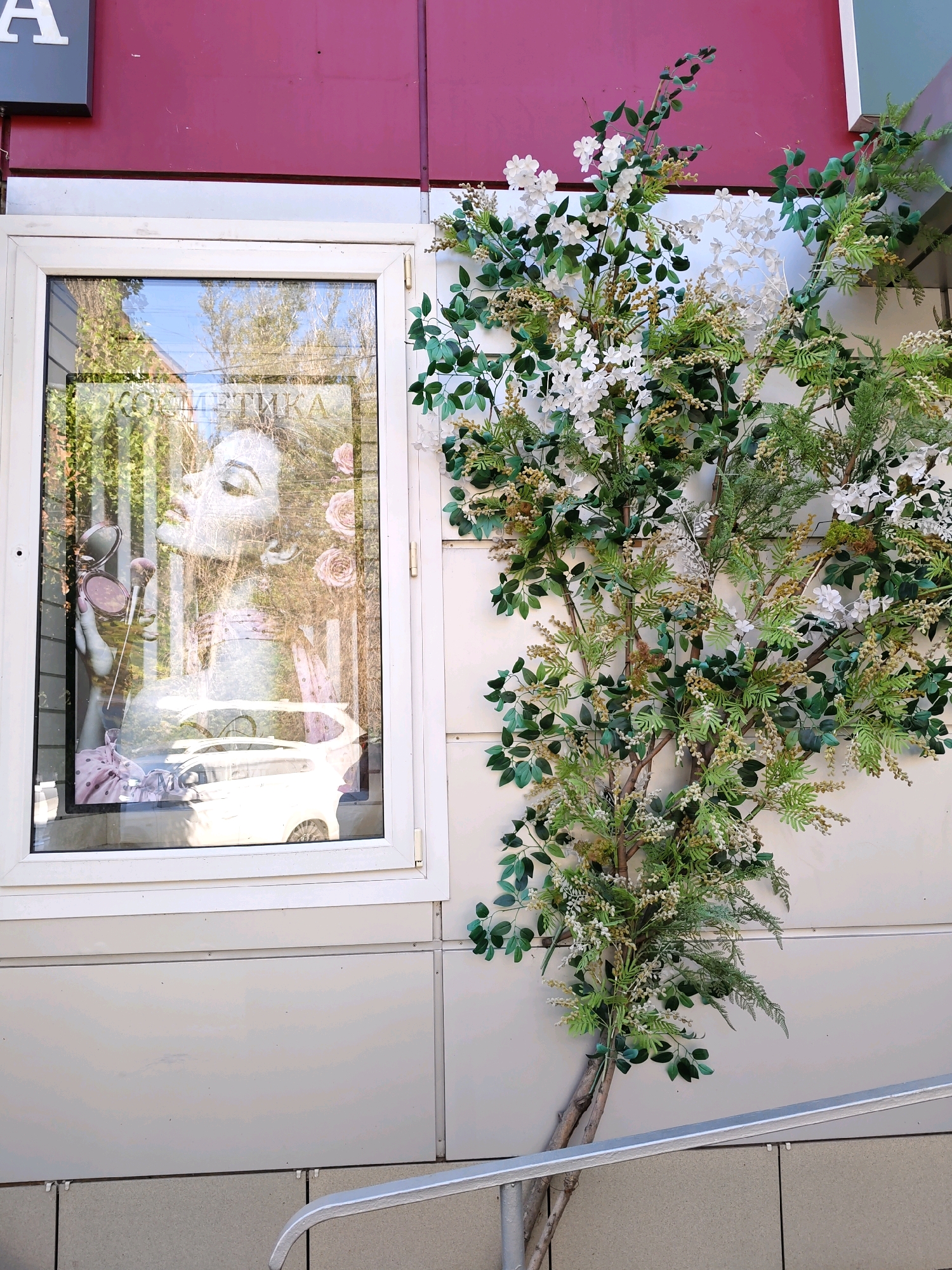 The image shows a storefront window with a decorative tree to its right. The storefront has a maroon-colored upper facade and a light-colored lower facade. The window is framed in white and displays a large image of a woman's face, with makeup brushes and a makeup compact, against a background of greenery and roses. The text "КОСМЕТИКА" (Cosmetics) is visible at the top of the display. The decorative tree is artificial, with green leaves and small white and beige flowers. The scene is set on a street in Rostov-on-Don, Russia. A metal railing is visible in the foreground.