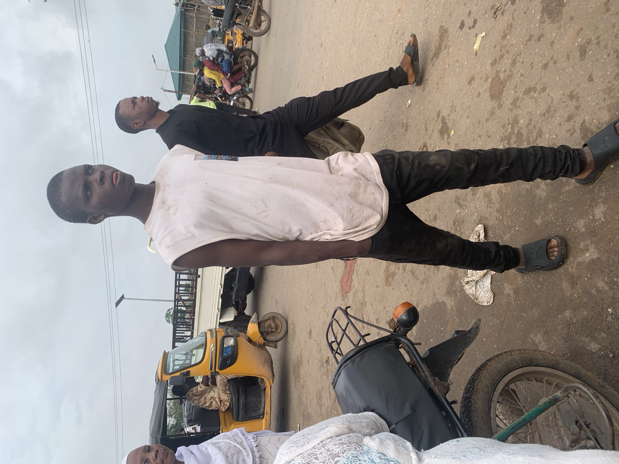 A medium shot shows two young Black men walking on a dirty, paved street in Shagamu, Nigeria. The man in the foreground is wearing a sleeveless white t-shirt and black pants, and he is looking to the right with his tongue sticking out. He is wearing black sandals. Behind him, another young Black man in a black long-sleeved shirt and black pants is walking in the same direction, also looking to the right. He is carrying a tan backpack over his left shoulder. In the background, a yellow motorized rickshaw, known as an "Okada," is parked. A man in a traditional tan robe is sitting in the driver's seat. Several other people and motorcycles are visible in the distance. The sky is overcast. The ground is rough and appears to be stained with dirt and possibly water.