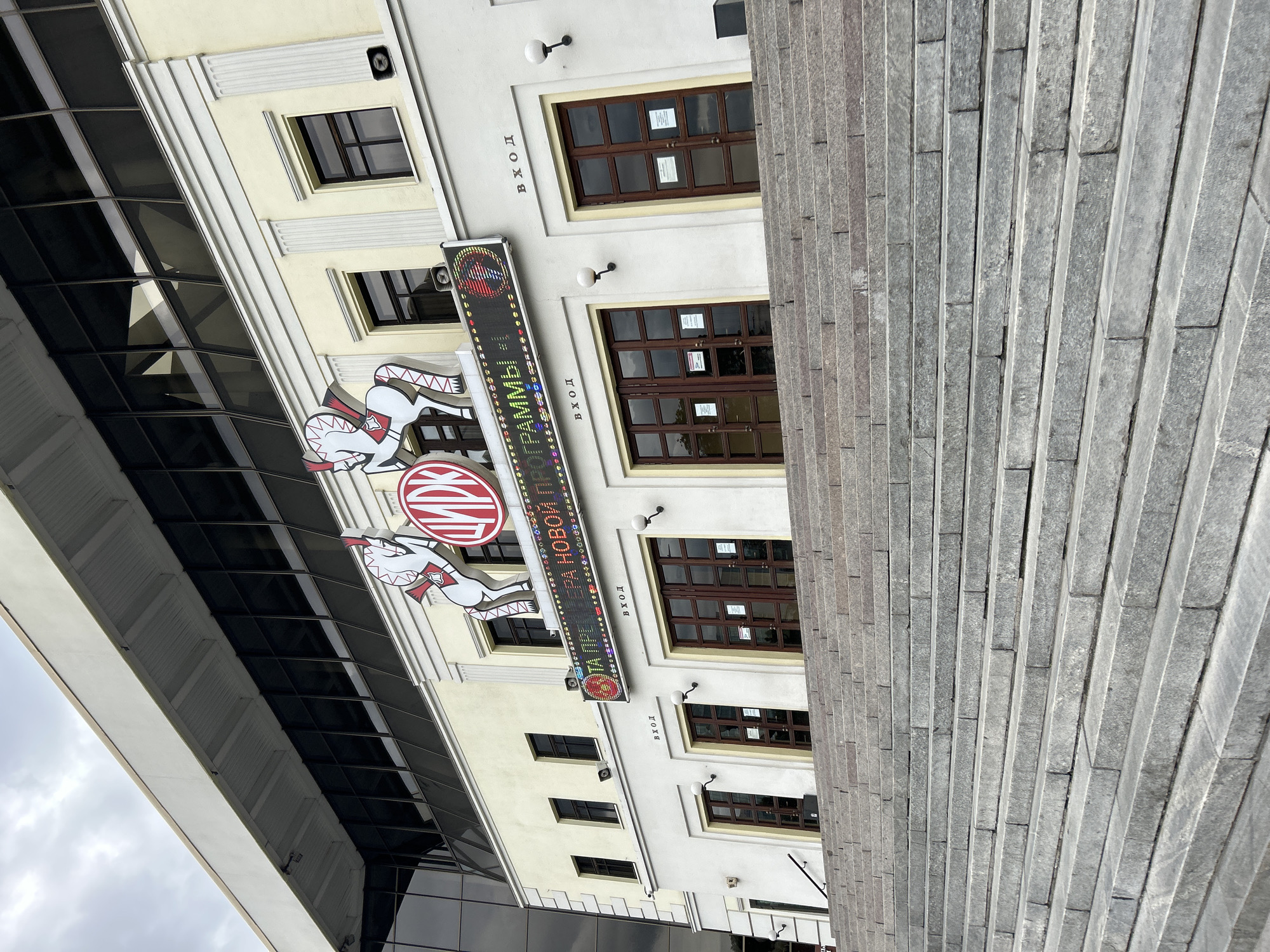 This image shows the exterior of the Moscow Circus on Tsvetnoy Boulevard. A wide, imposing set of stone stairs leads up to the building's entrance. The building itself is a light yellow color with a modern, darker glass-enclosed upper level. Above the entrance doors, a large marquee displays an animated message in Russian and colorful graphics, including stylized horses. The marquee reads "ЦИРК" (Circus) and "ТА ПРЕМЬЕРА НОВОЙ ПРОГРАММЫ" (New Program Premiere). Two wooden doors with glass panes are visible on the ground level, each marked with "ВХОД" (Entrance). The sky above is overcast.