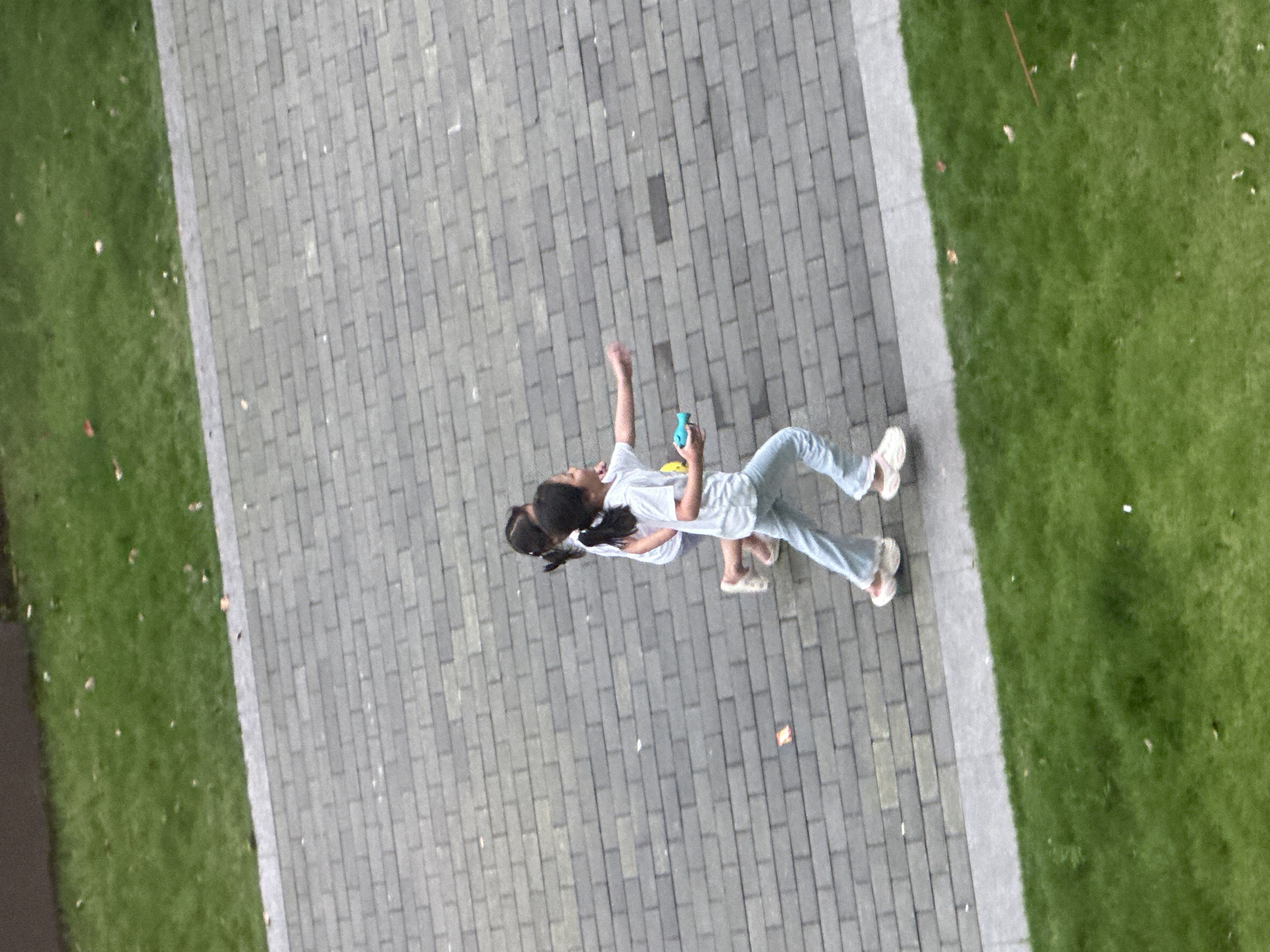 Two young girls are running side-by-side on a gray brick pathway, which is bordered by a strip of green grass on the left and possibly a dark-colored building or wall on the top. The girl in front is wearing a white t-shirt and light blue pants, and she is holding a small blue object in her right hand and appears to be reaching forward. The second girl, slightly behind and to the left, is wearing a white t-shirt and possibly light-colored shorts or a skirt, and her left arm is bent at the elbow as she runs. Both girls have dark hair tied back in ponytails. The lighting suggests it is daytime, and the scene is viewed from an elevated perspective looking down.