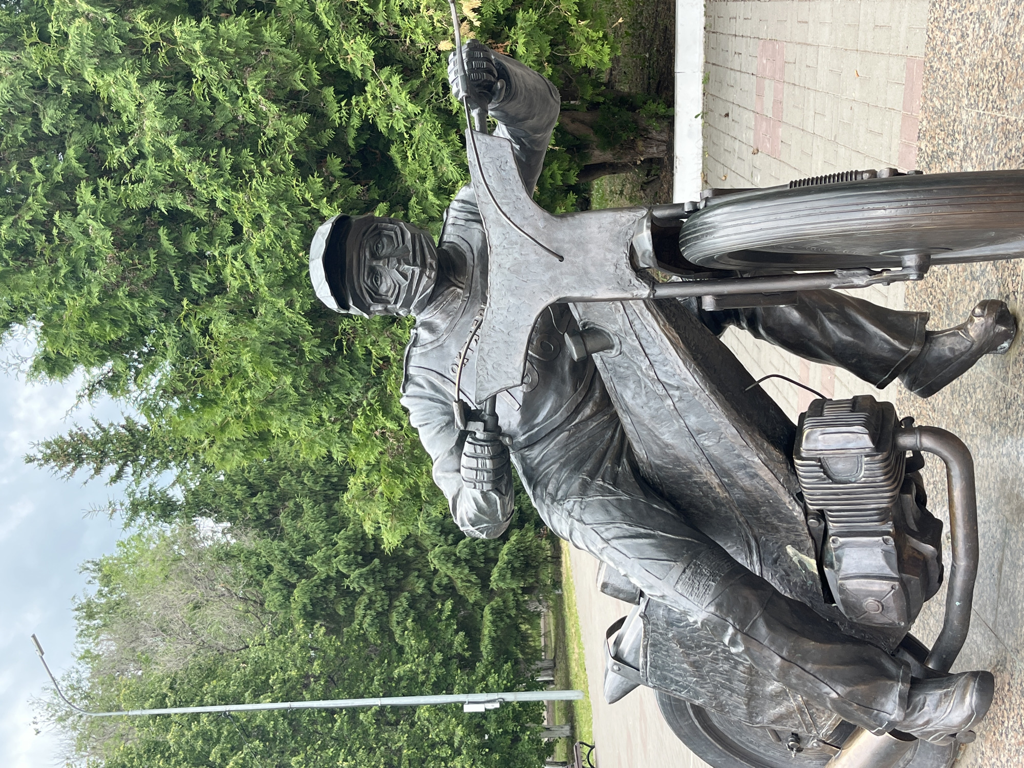 An outdoor, eye-level shot, rotated 90 degrees counter-clockwise, displays a detailed bronze statue of a motorcyclist and their motorcycle. The statue captures a dynamic moment, with the rider leaning forward as if in motion or making a turn.
The primary subject is a male figure, depicted in full racing gear, including a helmet covering his face with only the eyes visible through a visor-like opening. He wears a full-body racing suit with visible folds and detailing, and racing boots. His hands are firmly gripping the handlebars of the motorcycle, suggesting control and speed.
The motorcycle itself is intricately rendered in bronze, showcasing an exposed engine block with cooling fins, exhaust pipes, and a front wheel with a ribbed tire pattern. The overall design suggests a vintage or classic racing motorcycle.
The statue is positioned on a paved pathway, which features a light-colored, brick-patterned surface. Behind the statue, a dense backdrop of lush green trees with small, scale-like leaves (possibly conifer or thuja) fills the upper and left parts of the frame, indicating a park or landscaped area. A light pole or street lamp fixture is visible in the upper left background, against a bright, possibly overcast sky.
The scene takes place during daylight hours. The bronze material gives the statue a dark, metallic luster against the vibrant green of the foliage and the lighter pavement. No visible text is present in the image. The setting, in Oktyabrsky, Russia, suggests this might be a monument or public art installation celebrating motorcycling or a specific rider.
