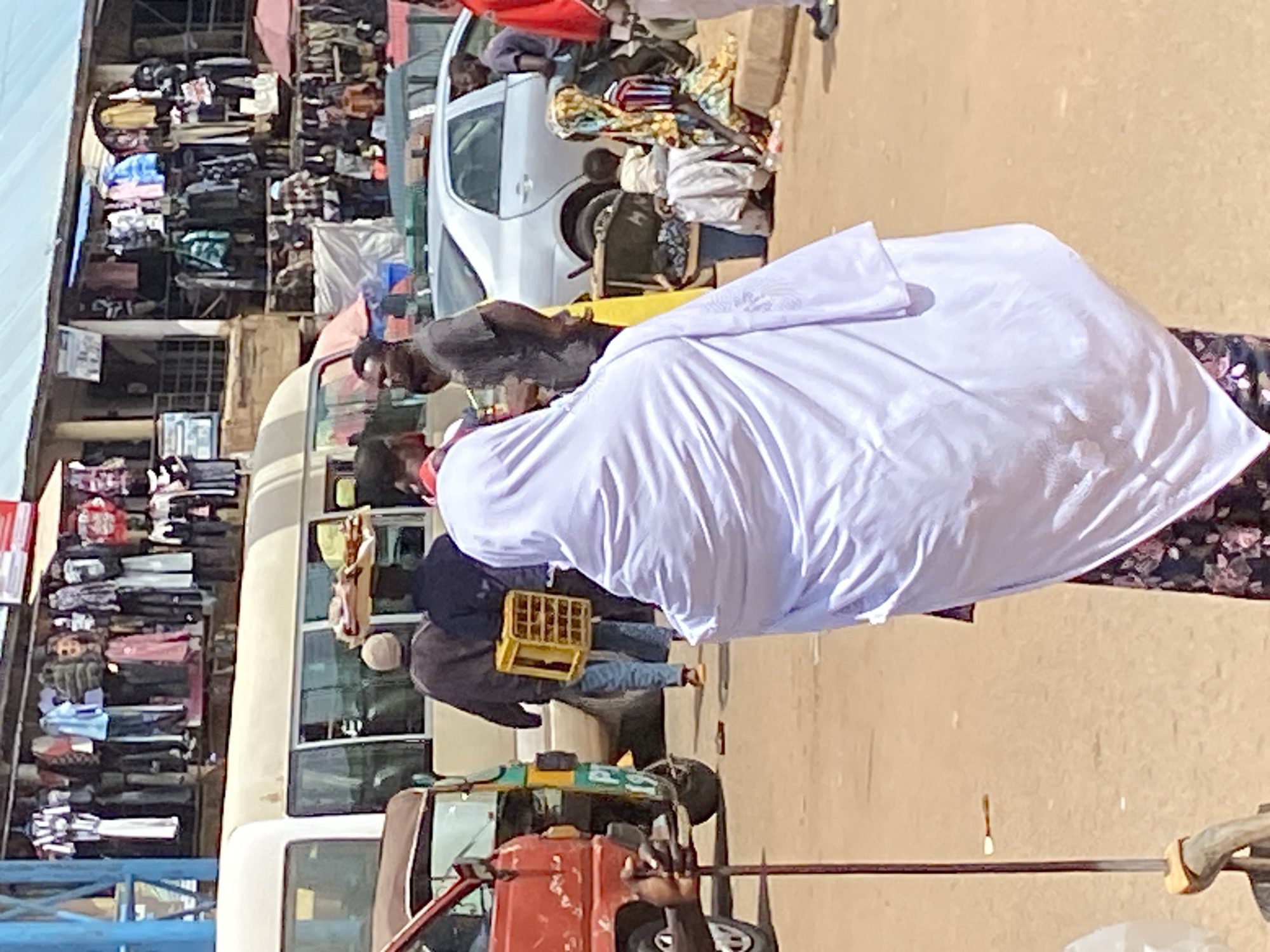 A crowded street market in Bukuru, Nigeria. A person in a white hijab is walking, carrying a yellow crate. Behind them, a white van is parked, and a red truck is visible in the foreground. Stalls filled with clothing and various items are set up along the street. The scene appears to be from daytime, with sunlight casting shadows. The atmosphere seems busy, typical of a bustling market.