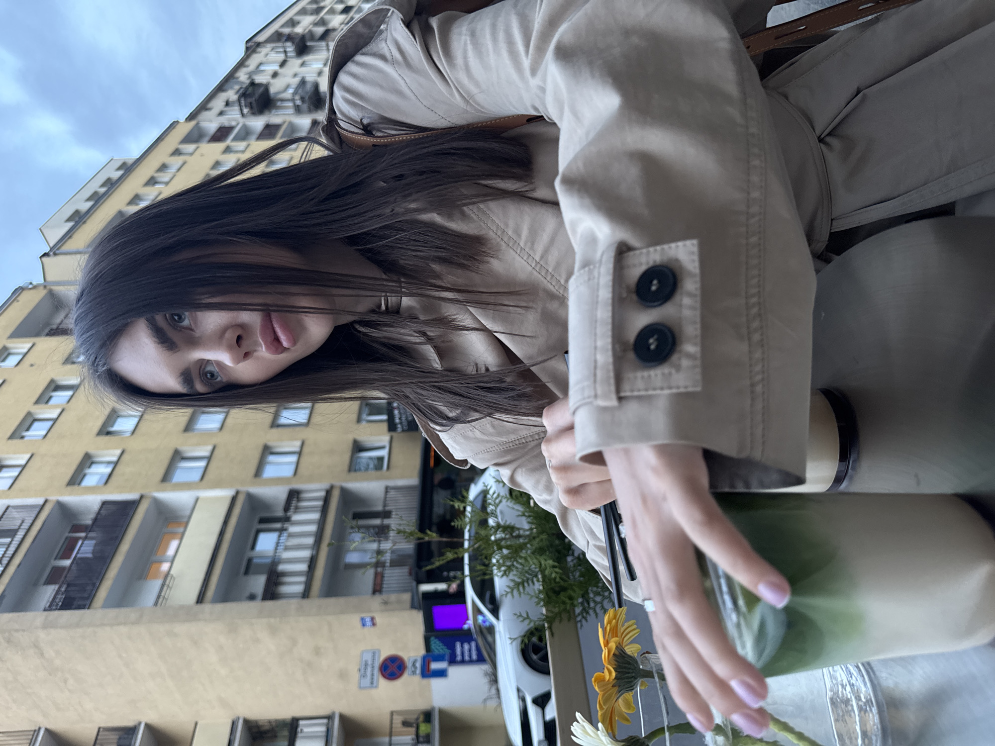 A young woman with dark hair and light eyes poses for a selfie at an outdoor cafe in Warsaw, Poland. She wears a light tan trench coat. The city street is behind her. She is holding a tall glass filled with a green-and-white liquid, and a vase with daisies sits in front of the glass. Apartment buildings are visible across the street. Street signs and a white car are also visible in the background.
