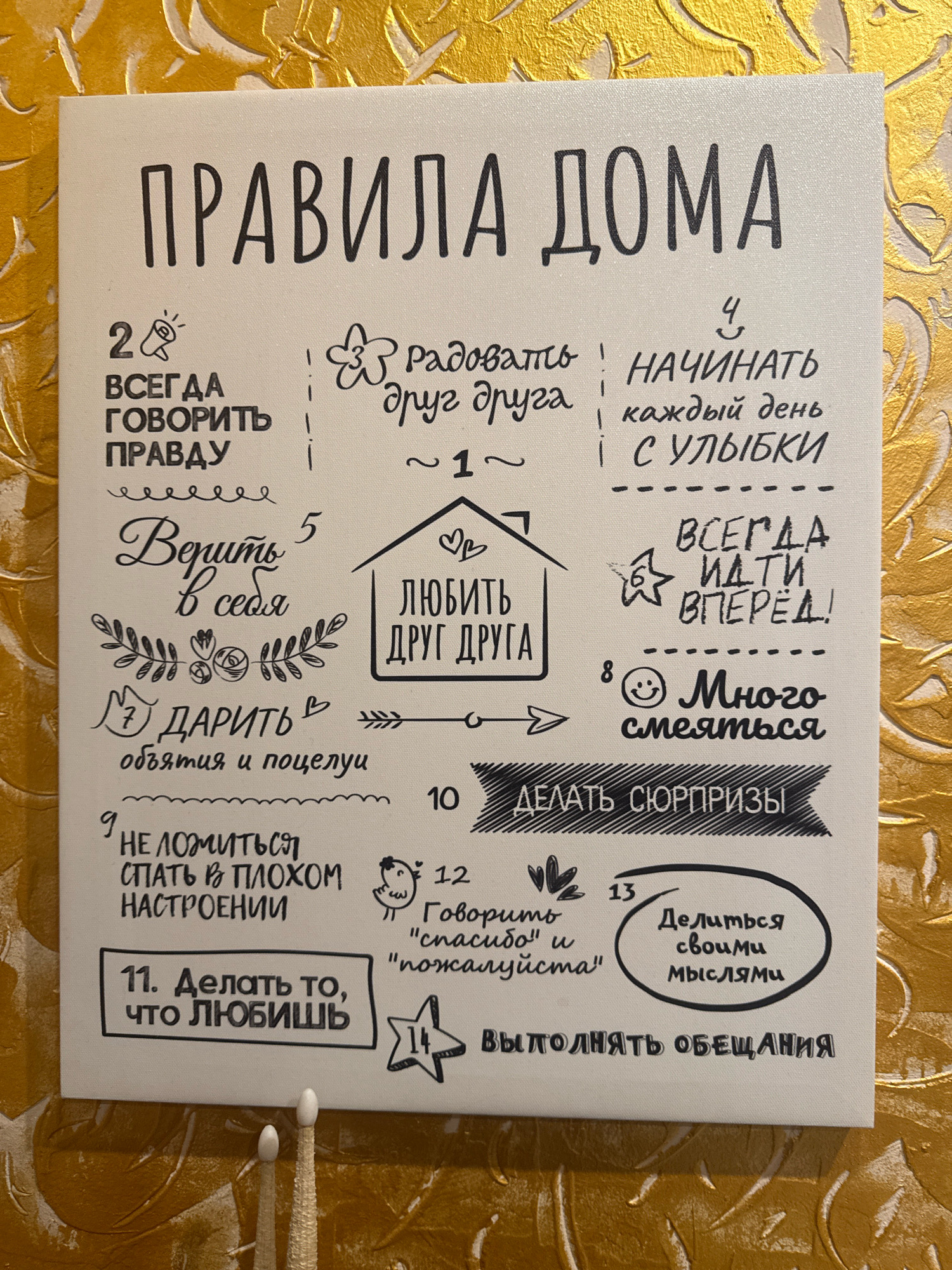 The image shows a canvas sign with "ПРАВИЛА ДОМА" (House Rules) written at the top in large, bold, black font. Below this title, there are numbered rules written in black text with accompanying illustrations. The sign is mounted on a textured, golden wall.
Here's a breakdown of the visible rules and their associated imagery:
* **2. ВСЕГДА ГОВОРИТЬ ПРАВДУ** (Always Tell the Truth): Illustrated with a stylized megaphone.
* **3. радовать друг друга** (Make Each Other Happy): Illustrated with a flower and a butterfly. Below this, there is a line with "~ 1 ~".
* **4. НАЧИНАТЬ КАЖДЫЙ ДЕНЬ С УЛЫБКИ** (Start Every Day With a Smile): Illustrated with a smiley face.
* **5. Верить в себя** (Believe in Yourself): Written in a more cursive font and accompanied by a decorative floral border.
* **ЛЮБИТЬ ДРУГ ДРУГА** (Love Each Other): Written inside a house silhouette.
* **6. ВСЕГДА ИДТИ ВПЕРЁД!** (Always Move Forward!): Illustrated with a star.
* **7. ДАРИТЬ объятия и поцелуи** (Give Hugs and Kisses): Illustrated with a hand presenting a heart and small hearts.
* **8. Много смеяться** (Laugh a Lot): Illustrated with a smiley face.
* **9. НЕ ЛОЖИТЬСЯ СПАТЬ В ПЛОХОМ НАСТРОЕНИИ** (Don't Go to Bed in a Bad Mood): Illustrated with scribbled lines.
* **10. ДЕЛАТЬ СЮРПРИЗЫ** (Make Surprises): Written on a banner-like graphic with stripes.
* **11. Делать то, что любишь** (Do What You Love): Enclosed in a rectangular box.
* **12. Говорить "спасибо" и "пожалуйста"** (Say "Thank You" and "Please"): Illustrated with a small bird.
* **13. Делиться своими мыслями** (Share Your Thoughts): Enclosed in a circular outline.
* **14. ВЫПОЛНЯТЬ ОБЕЩАНИЯ** (Keep Promises): Illustrated with a star.
The lighting is slightly warm, suggesting an indoor setting. The golden textured wall indicates a decorative interior. The overall impression is of a warm and encouraging environment, possibly a family home.
The image does not contain any specific visual cues that would allow for precise identification of its location within the city of Almaty, Kazakhstan. It depicts a common decorative item found in homes worldwide.