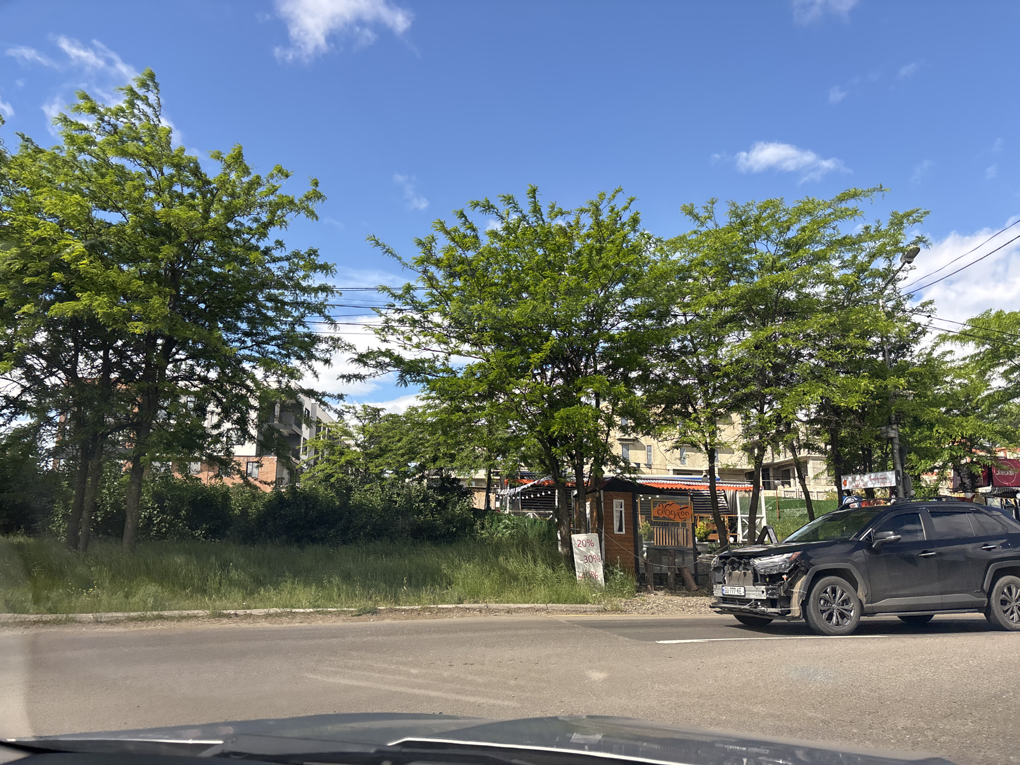 The image depicts a street scene in Tbilisi, Georgia. A black Toyota RAV4 with front-end damage is parked on the right side of the road, partially obstructing a white lane marker. The license plate number is GA-777-KE. Behind the car, there is a small wooden structure, possibly a shop or stall. In front of the building, there is a sign with the numbers "20%" and "30%". Buildings are visible in the background, obscured by green trees. The sky is blue with scattered white clouds. The photograph was taken from inside another vehicle, with the dashboard visible at the bottom.