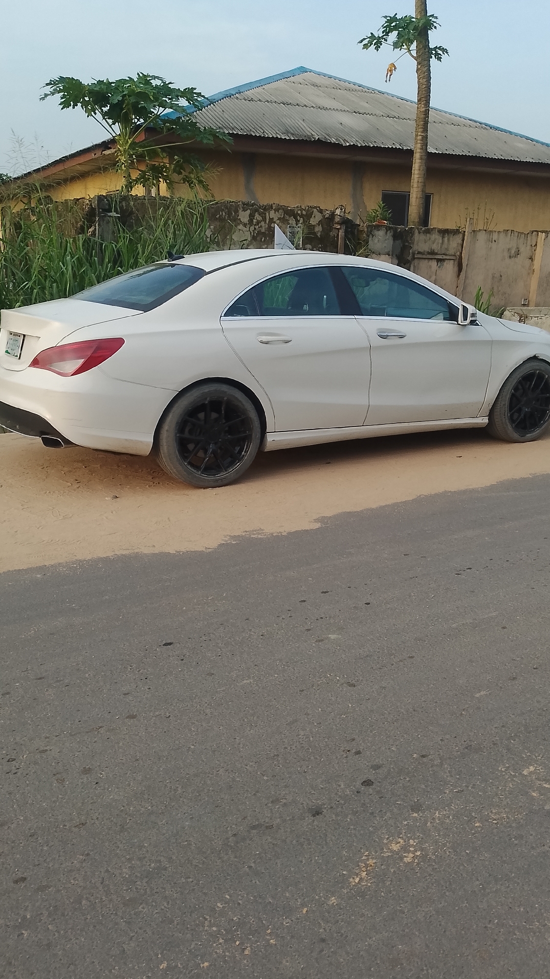 A white Mercedes-Benz CLA is parked on the side of a paved road, with the front end facing away from the viewer. The car has black rims. The background includes a building with a blue corrugated metal roof and vegetation. The ground adjacent to the road is dirt and sand. The location appears to be Asubi, Nigeria.