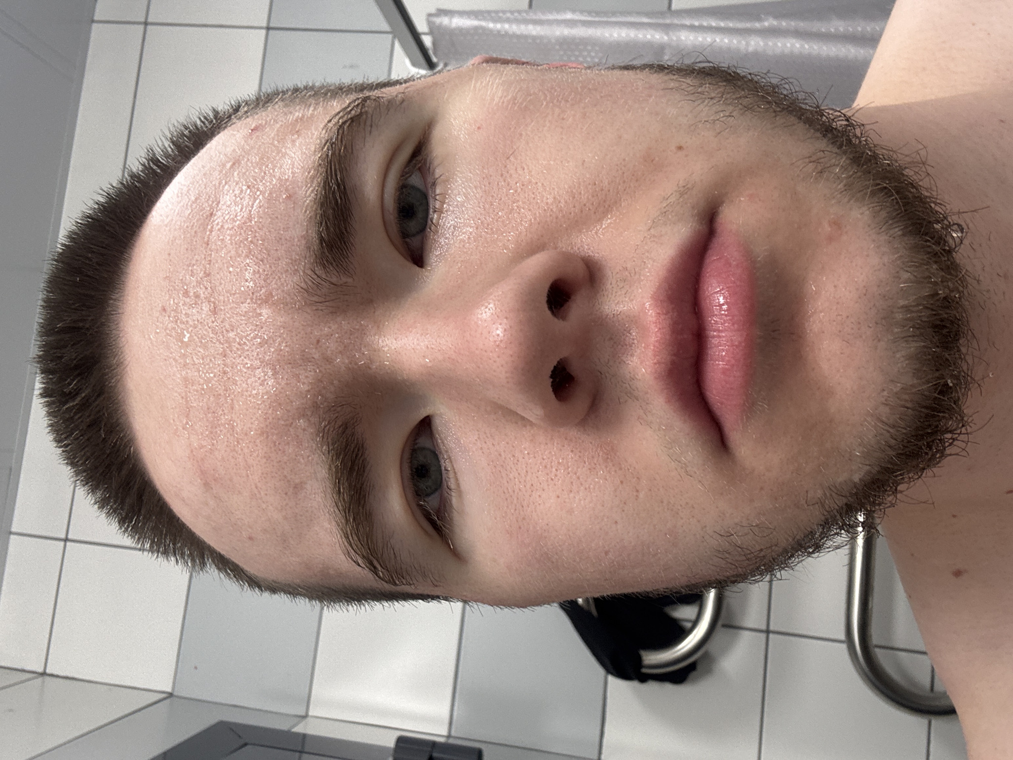 The image is a close-up, slightly angled portrait of a young male, likely in his late teens or early twenties. He has short, dark brown hair, dark eyebrows, and light-colored eyes. A light brown beard and stubble frame his jawline and chin. His skin appears flushed and damp with sweat, particularly noticeable on his forehead, nose, and cheeks, suggesting recent physical activity or a warm, humid environment. His lips are full and slightly parted.
The setting appears to be an indoor, utilitarian space, possibly a bathroom, shower room, or locker room. The background features white tiled walls with visible grout lines. In the upper background, a translucent shower curtain or partition is partially visible. To the lower right, a metallic, U-shaped grab bar or towel rack is present, with a dark piece of fabric partially visible beneath it. The subject's expression is neutral, possibly conveying a sense of calm or tiredness. There is no visible text within the image. While the provided context specifies the location as Kar'yer, Russia, there are no discernible visual cues within the photograph to corroborate this geographic detail.