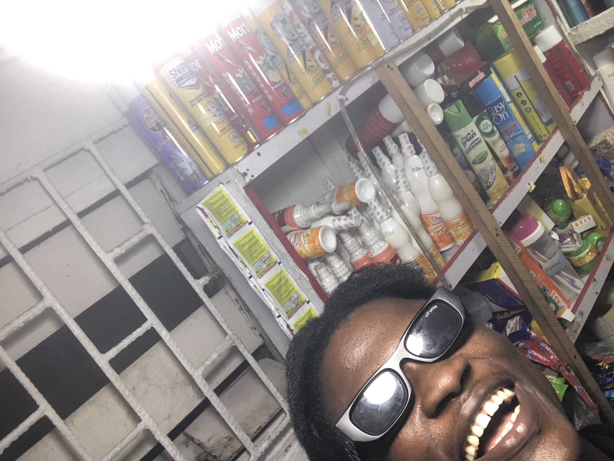 The image shows a close-up of a person wearing black sunglasses and smiling broadly, with an out-of-focus background of a store shelf filled with various products, including aerosol cans and bottles of what appear to be cleaning supplies or personal care items. The store is likely located in Ado, Nigeria, as indicated by the context. The shelves are made of wood and the products are arranged in a somewhat cluttered manner. The person's face takes up a significant portion of the foreground, with their teeth clearly visible in their open mouth. The lighting suggests an indoor environment, possibly a small retail shop.