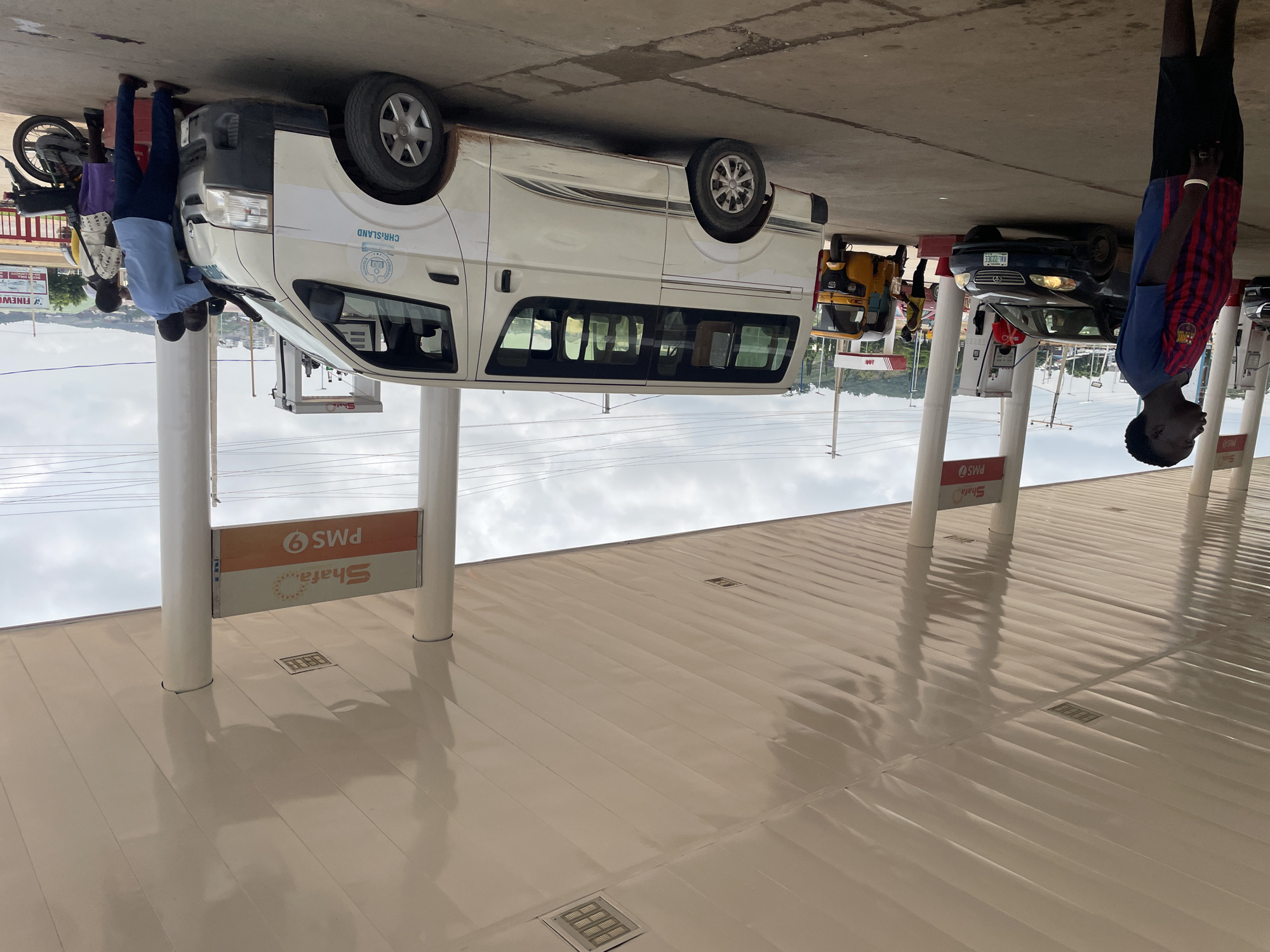 The image depicts a scene at a Shafa gas station in Abuja, Nigeria. Several vehicles are present, including a white van, a dark sedan, and a yellow tricycle. People are interacting with the vehicles, with some appearing to be refueling or getting ready to refuel. The gas station has a covered canopy supported by white pillars, and visible signage indicates "Shafa PMS 9." The environment appears to be an outdoor setting with a paved ground surface and a cloudy sky. The overall impression is a functional, everyday scene at a Nigerian fuel station.