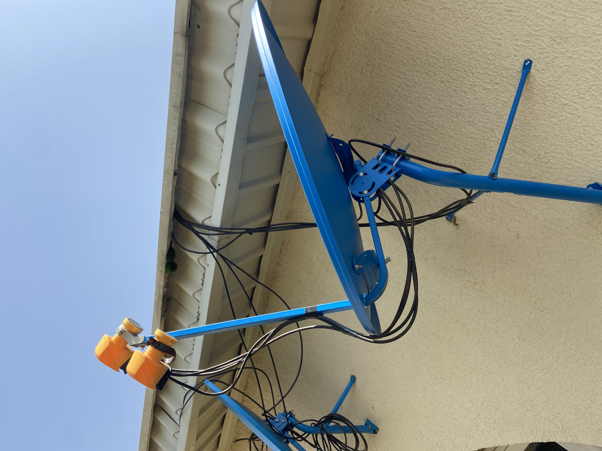 The image shows a close-up, low-angle shot of a blue satellite dish mounted on a stucco wall. The dish is tilted upwards, with its reflective surface prominently displayed. Two orange LNBs (Low-Noise Block downconverters) are attached to the arm of the dish. Several black cables are bundled and run from the LNBs, down the arm, and towards the wall.
The setting appears to be outdoors, with a clear blue sky visible in the upper left portion of the image. A portion of a white, corrugated roof overhang is visible on the left side, partially obscuring the sky. The wall is textured stucco, a common building material.
There are no people visible in the image. The activity taking place is the installation or presence of the satellite dish, likely for receiving television or internet signals.
Notable details include the bright, clear sky suggesting it is daytime, likely sunny. The angle of the dish suggests it is pointed towards a satellite in orbit. The stucco texture of the wall and the visible roof suggest a residential or commercial building. There is no visible text in the image. The overall impression is of functional outdoor equipment against a bright sky and textured wall.