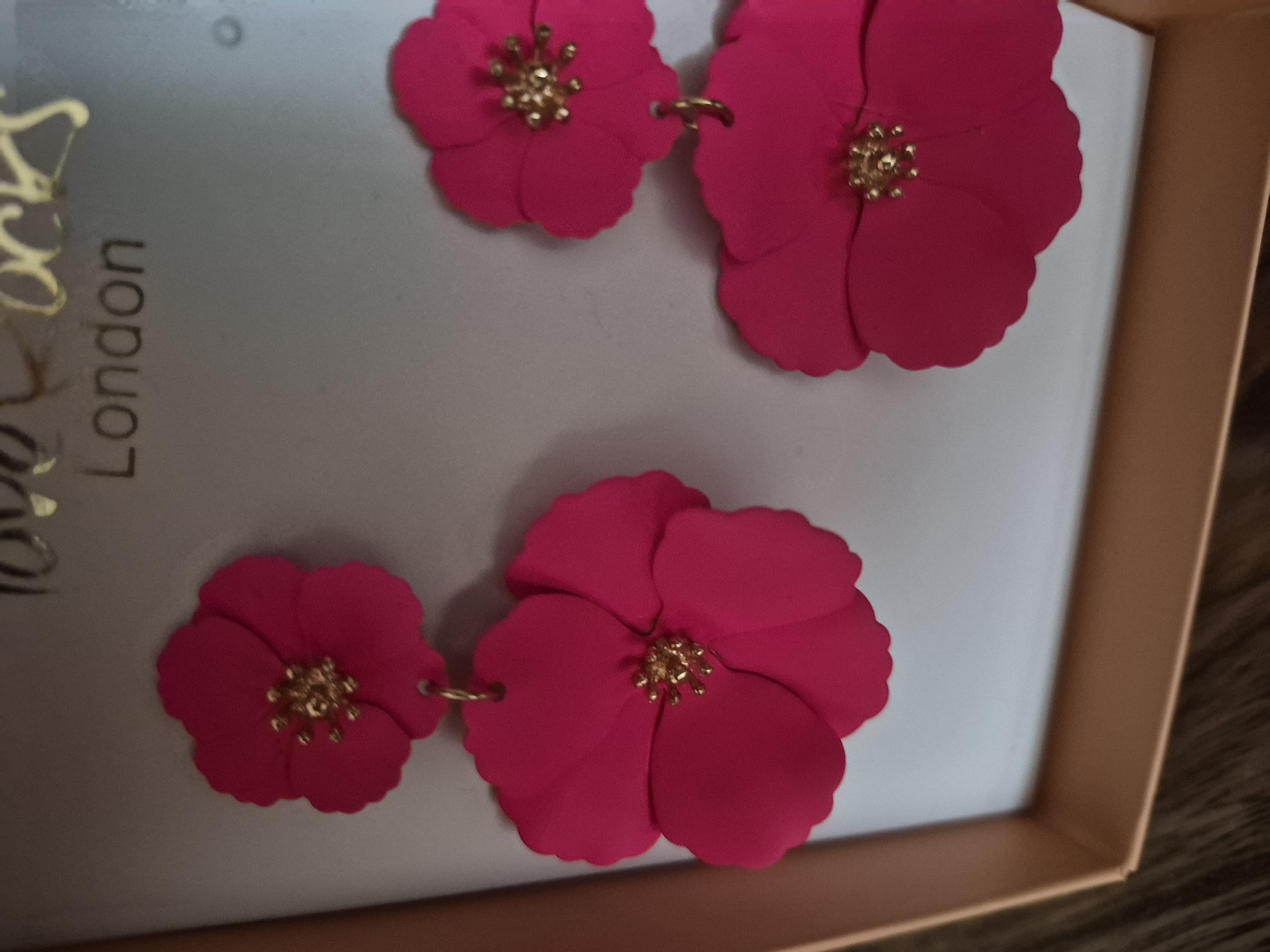The image shows a pair of fuchsia pink, flower-shaped drop earrings against a grey background. Each earring features a large, multi-petaled flower with a gold-colored beaded center. The earrings are hanging from what appears to be packaging or a display box, with the brand name "London" partially visible at the top of the background.