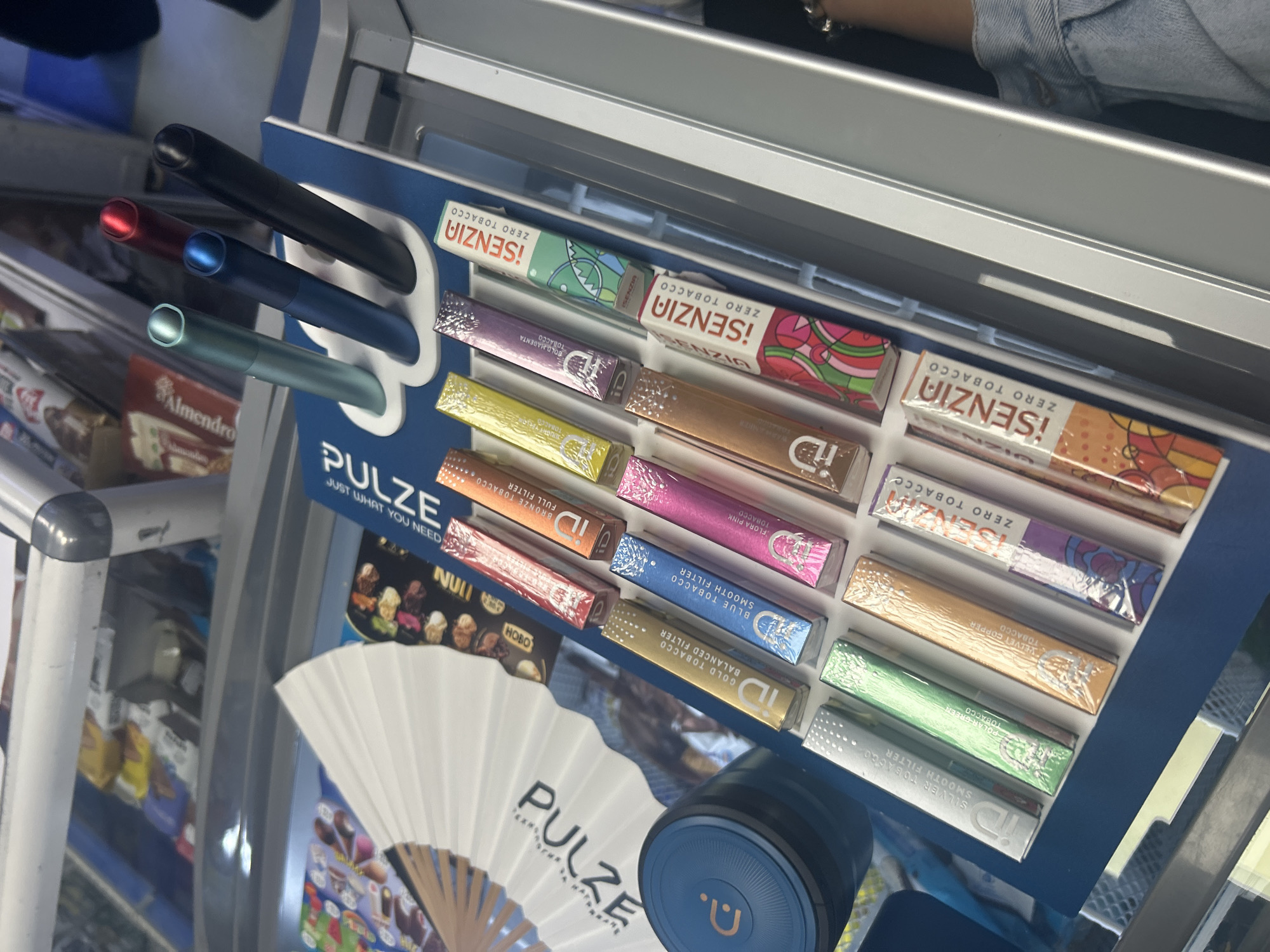The image shows a close-up, eye-level, outdoor shot of a retail display. The display features an array of vaping products, including multiple "iQOS" branded tobacco sticks in various colors and flavors, such as "Gold Tobacco," "Blue Tobacco," "Flora Pink Tobacco," "Silver Tobacco," "Polar Green Tobacco," and "Velvet Copper Tobacco." There are also several "¡SENZIA ZERO TOBACCO" sticks in colorful packaging. In the upper part of the display, three vaping devices are visible in teal, dark blue, and black, with a red-tipped device behind them. A white fan with black lettering that says "PULZE" is positioned to the left of the tobacco sticks. A dark blue cylindrical container with an orange logo is also present on the left side of the frame. The background suggests a store environment with shelves of products, including some visible in the upper portion of the image. The overall lighting is bright, indicating an indoor setting or a shaded outdoor area.