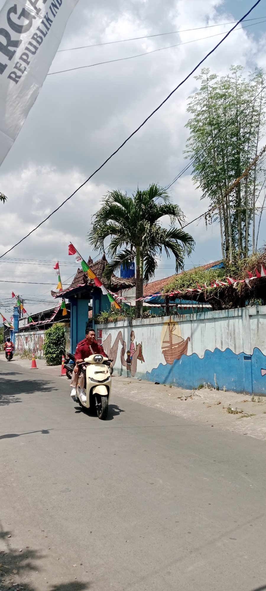 The image captures a street scene in Purwomartani, Indonesia, on a partly cloudy day. A man wearing a red shirt and tan pants is riding a light-colored scooter down a paved road. Behind him, another scooter is partially visible, also being ridden. The road is flanked by a low wall decorated with a mural of a ship on blue waves and a figure. Numerous colorful flags, some appearing to be Indonesian flags (red and white), are strung along the top of the wall and from a building on the left. A tall palm tree stands near the wall, and bamboo-like plants are visible in the background. Overhead, power lines crisscross the sky, and a white banner with dark text, partially obscured by the sky, is visible in the upper left quadrant. The overall environment suggests a festive or celebratory atmosphere, possibly related to Indonesian Independence Day.