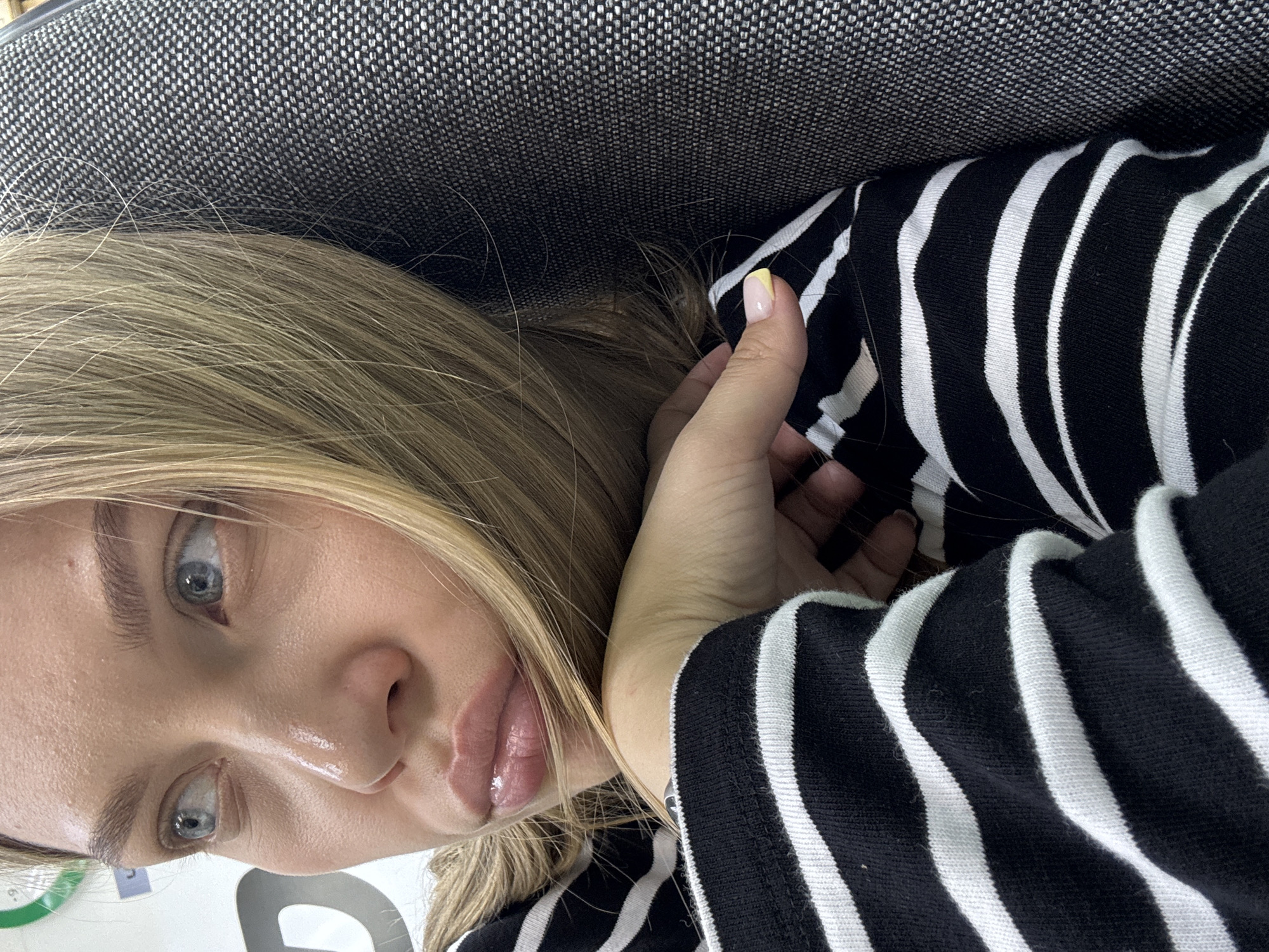 A close-up, slightly tilted portrait shows a young person, likely female, reclining indoors. The subject has long, light blonde hair, striking light blue or grey eyes, and full, slightly parted lips. Their skin appears dewy and moisturized, particularly around the nose and mouth.
They are wearing a long-sleeved top with prominent black and white horizontal stripes. One hand is visible, resting near their shoulder, with a fingernail showcasing a stylish design of a yellow tip over a clear or light pink base. The person is positioned against a textured dark gray or black upholstered surface, possibly a chair, sofa, or headrest. A few individual blonde hairs are visible against this dark background.
In the bottom left corner, indistinct shapes of white and green are visible, possibly a blurred logo or text on an object in the background, but no specific text is legible. The gaze is direct and engaging, suggesting a moment of rest or observation. The image provides no specific cues regarding the time of day or weather, nor does it contain explicit location markers for Chaikovsky, Russia, beyond the general indoor setting.