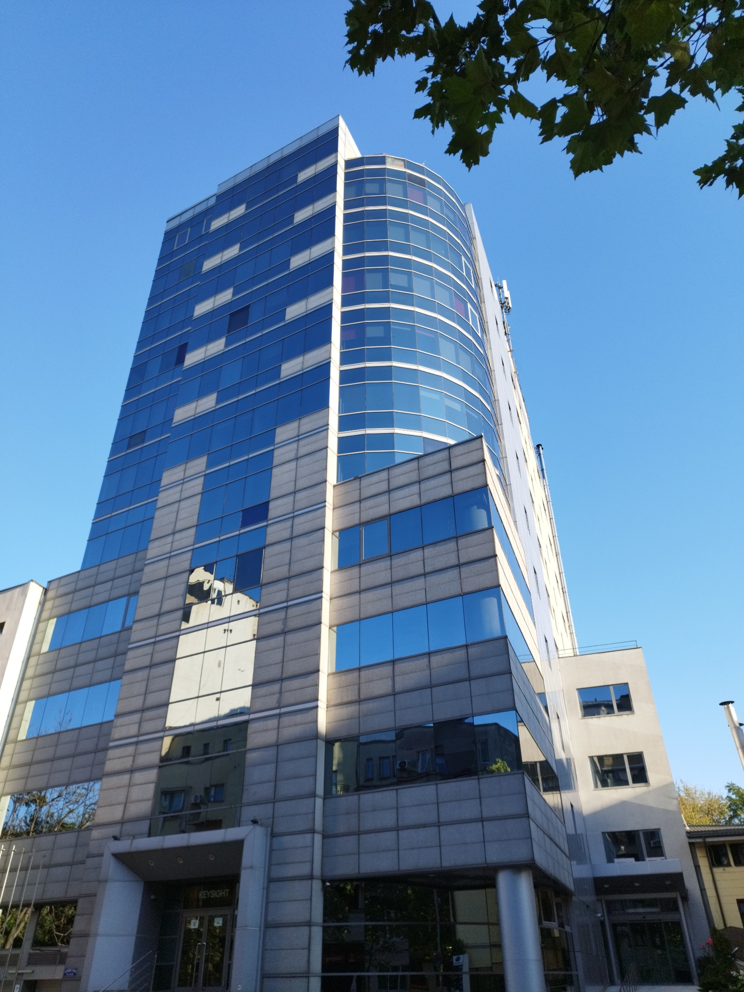 The image shows a modern glass-fronted office building in Bucharest, Romania, under a clear blue sky. The building is tall and features a distinctive curved facade with a pattern of blue and white glass panels on its upper section, and gray panels on its lower sections. Reflections of other buildings and trees are visible in the glass. The entrance to the building is visible at the bottom, with a sign that reads "KEYSIGHT". Some trees with green leaves are seen in the foreground, partially obscuring the top of the building. The building appears to be situated in an urban environment.