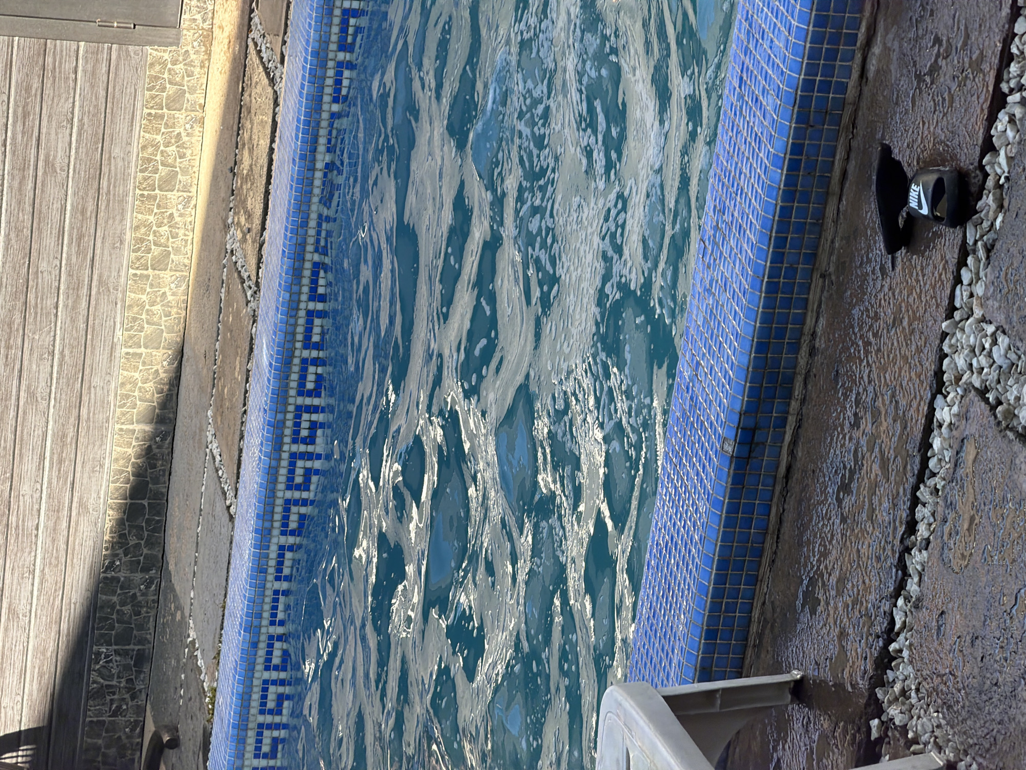 The image shows a close-up of the edge of a swimming pool, with the water rippling and reflecting sunlight. The pool is bordered by blue mosaic tiles. Beyond the pool, there is a wet, stone-paved area with a few white pebbles scattered around. In the background, a wooden fence and a stone wall are visible, casting shadows. On the wet ground near the edge of the pool, a pair of black Nike slides is placed. The scene appears to be outdoors, in a setting that could be a private residence or a public facility. The presence of a pool and slides suggests a recreational or leisure context. The city of Shymkent, Kazakhstan is not explicitly depicted or indicated within the visual elements of the image.