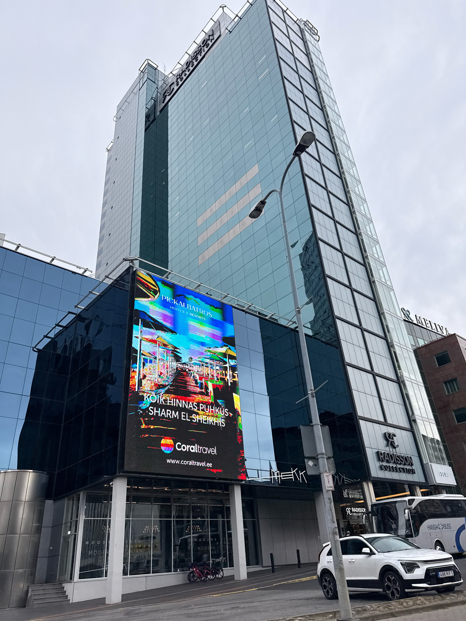 This is a street-level, low-angle shot of the Radisson Collection Hotel, Tallin, located in Tallinn, Estonia. The modern glass-and-steel skyscraper dominates the frame, its facade reflecting the overcast sky. A large, vibrant digital billboard is prominently displayed on the lower section of the building, advertising "PICKALBATROS HOTELS & RESORTS" and "Coral Travel" with an image of a beach resort. Below the billboard, the ground floor features large glass windows revealing a modern interior with text visible, possibly indicating a restaurant or shop. A white SUV and a bus are parked on the street in front of the building. Two bicycles are parked on the sidewalk near the entrance. The overall scene conveys a sense of urban architecture and commercial activity.