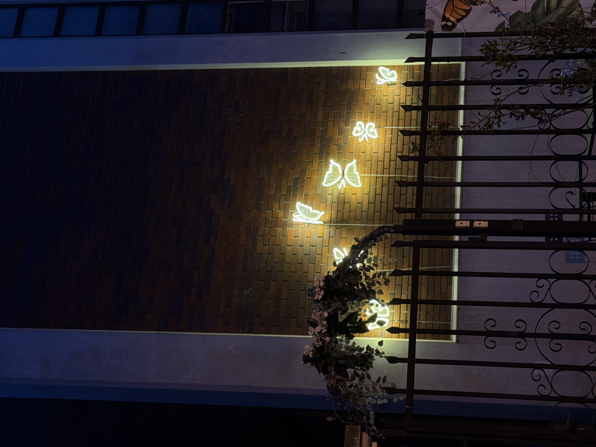 A nighttime image depicts a building facade in Varna, Bulgaria. The background features a brick wall, illuminated with an amber glow. Five luminous butterfly-shaped lights, emitting a bright yellow-white illumination, are mounted diagonally upwards on this wall. A decorative arch of artificial green foliage and pale flowers is draped over the top of a dark metal gate in the foreground. The gate consists of vertical bars with pointed finials and decorative scrollwork in its lower portion. To the right of the gate, a partial, large banner displaying an orange butterfly wing is visible. Two light-colored rectangular devices are affixed to a vertical surface next to the gate. Concrete pillars frame the illuminated brick section, with a multi-story building featuring dark windows partially visible on the far right. No individuals are present. The precise location within Varna is not identifiable from the image.