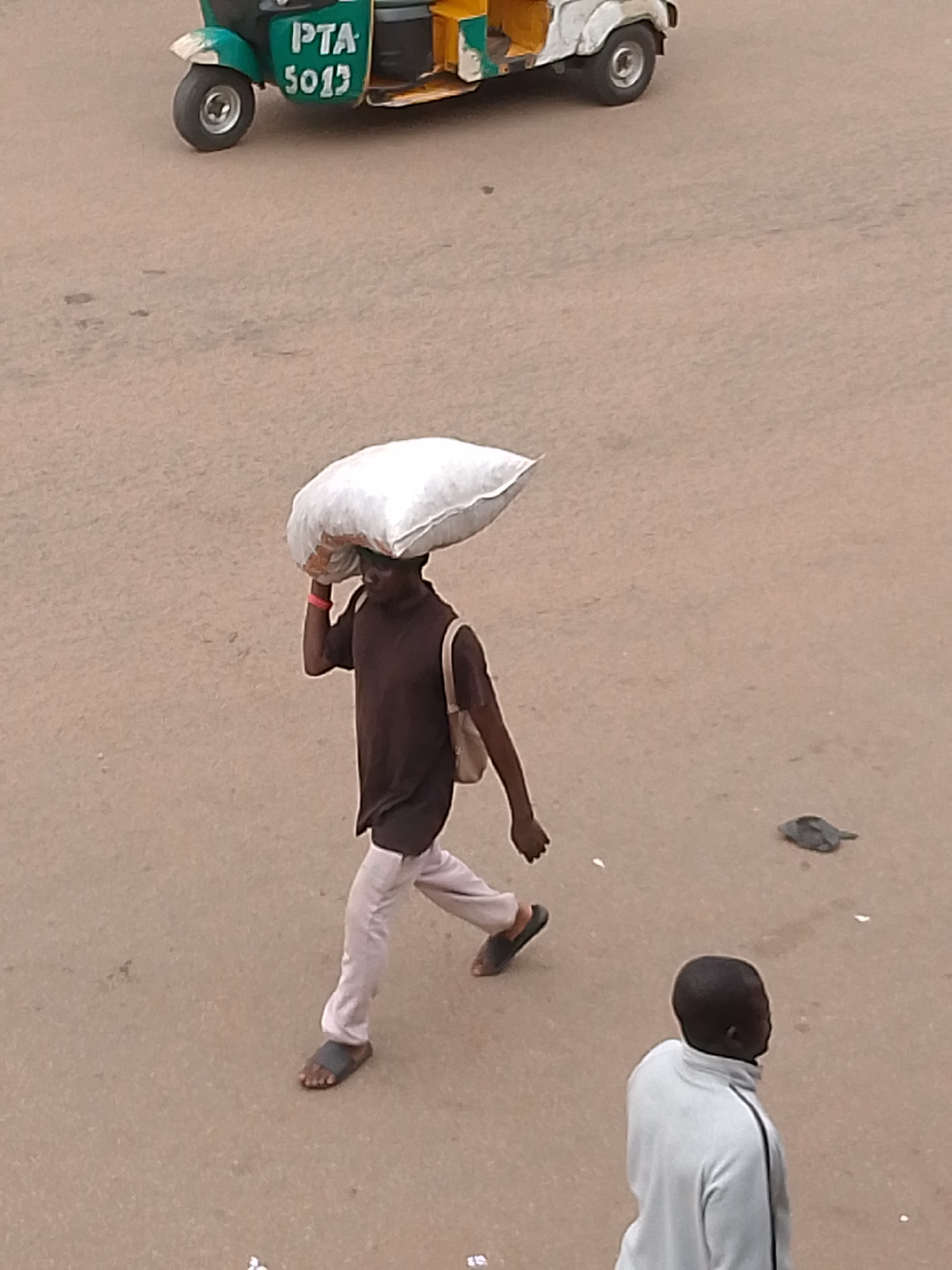 A man in a brown shirt and white pants walks on a dirt surface with a large white bag balanced on his head. He wears sandals. A second man, wearing a white jacket, stands with his back to the viewer. In the top portion of the frame is a green and white motorized tricycle with "PTA 5013" painted on the side. This image was taken in Bukuru, Nigeria.
