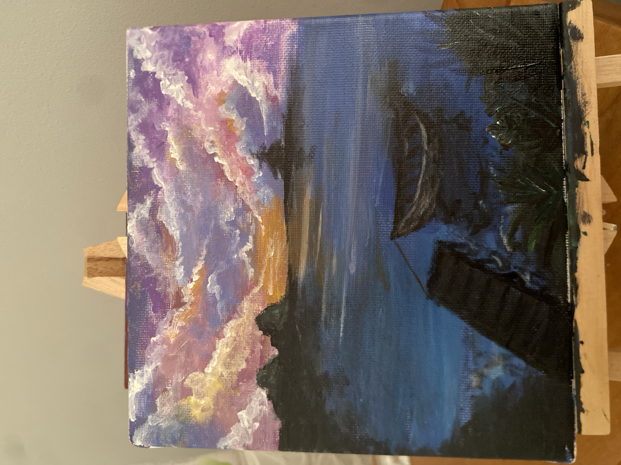 The image shows a square-format oil painting on canvas, resting on a wooden easel. The painting depicts a landscape with a colorful sunset or sunrise sky transitioning into a dark, stylized rendering of water and vegetation, possibly a lake scene with trees and a building. The left portion of the painting features clouds painted with shades of purple, pink, white, and hints of yellow. The right portion is dominated by dark blue and black, representing water and trees, with a geometric shape suggesting a building or structure reflected in the water. The setting is not identifiable as North Kuta, Indonesia from the image alone.
