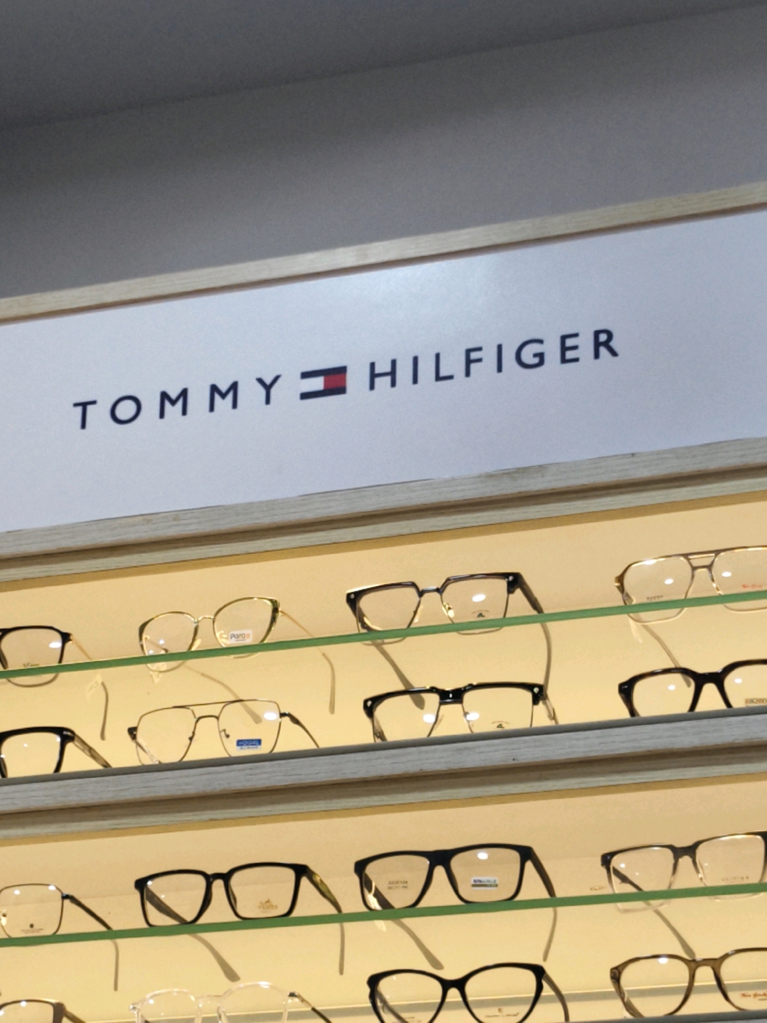 The image displays a row of eyeglasses on shelves in what appears to be an optical shop. A sign above the shelves reads "TOMMY HILFIGER." The eyeglasses are of various styles and frame colors, including black, gold, and clear. The background is a plain, light-colored wall. The lighting is bright and even, illuminating the products. The image appears to be taken from a medium distance, focusing on the display of glasses and the brand signage. The location "Modhera Char Rasta, India" is stated as context, suggesting this is a commercial setting in that area.