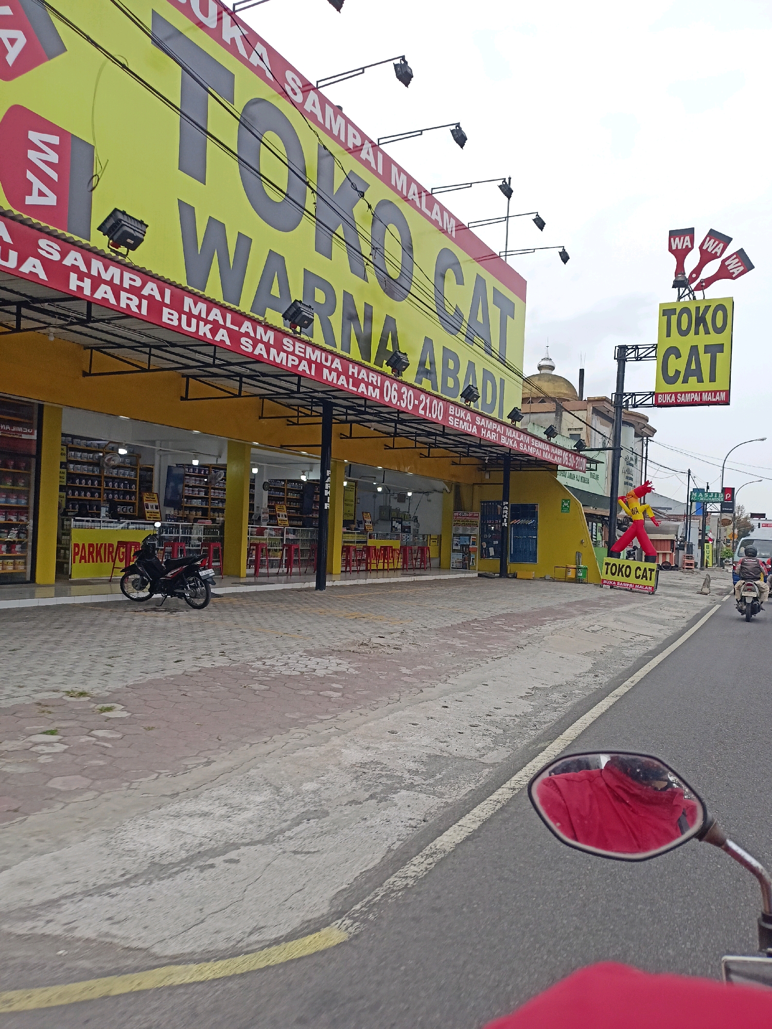 The image depicts a street view in Magelang, Indonesia, with a prominent yellow building labeled "TOKO CAT WARNA ABADI" (Eternal Color Paint Store). The store has a large yellow banner with red lettering, advertising paint products. There are also smaller signs with "WA" (WhatsApp) indicating contact information. The store appears to sell a variety of paints, with shelves of cans visible through the open storefront. A red motorcycle is parked in front of the store. Further down the street, there's a bright yellow inflatable figure dressed in red and yellow, also advertising "TOKO CAT" (Paint Store). Traffic is visible on the right side of the road, including motorcycles and a car. The sky is overcast. The overall scene captures a commercial area with businesses and a street in Indonesia.