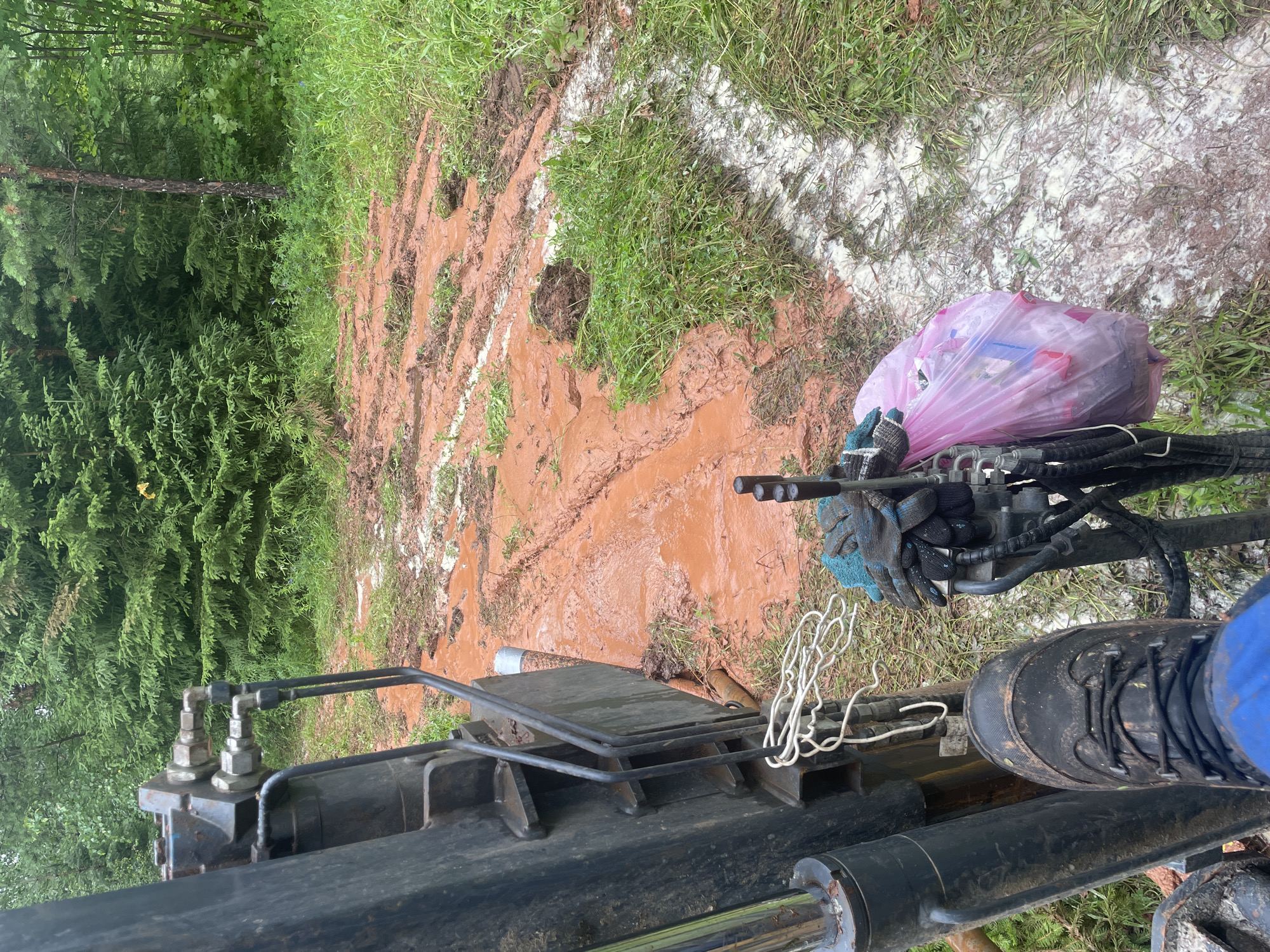 The image depicts a muddy, outdoor scene with a heavy-duty machine in the foreground. The machine appears to be a piece of construction or drilling equipment, with visible hydraulic components, hoses, and a metal arm. A person's black boot, covered in mud, is placed on one of the machine's parts, suggesting an operator is present. They are wearing blue work pants. On the machine, a pink plastic bag filled with what seems to be trash is tied to a piece of equipment. Beside the bag, a pair of dark gloves with blue accents are resting.
The ground is heavily marked with tire tracks, indicating recent activity by heavy vehicles. The mud is a reddish-brown color, suggesting iron-rich soil. Patches of white material are scattered across the ground, possibly construction debris or a substance used in the work. The background features lush green foliage, including coniferous trees and grass, indicating a natural or semi-natural environment.
The overall impression is one of ongoing work in a challenging, muddy terrain, possibly in a rural or forested area. The weather appears to be overcast or the lighting is diffused, as there are no strong shadows. The scene is static, with no clear indication of time of day, other than it is daytime. The presence of the trash bag suggests some form of site work or cleanup. No visible text is present in the image.