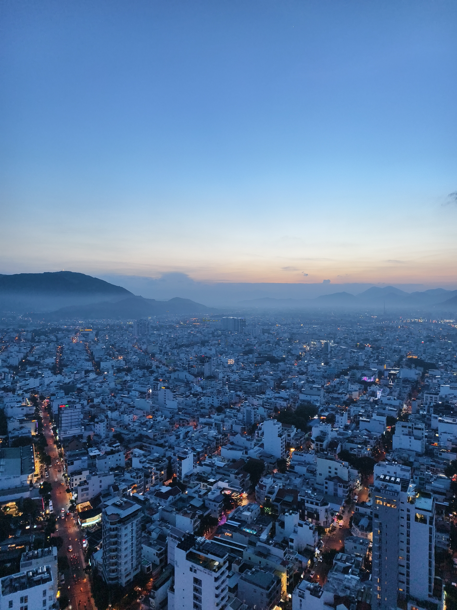 This detailed image captures an expansive high-angle view of a dense urban area during twilight, likely early evening, in Vung Tau, Vietnam.
The upper third of the frame is dominated by a clear sky, transitioning from a deep indigo blue at the top to a lighter, paler blue and faint orange glow near the horizon, indicative of a recent sunset. A single bright celestial object, possibly a star or planet, is visible in the upper left quadrant.
In the mid-ground, a range of dark, hazy mountains forms a prominent backdrop, silhouetted against the fading light of the horizon. On the peak of the leftmost and most defined mountain, a large, illuminated white statue, resembling a Christ the King figure, stands distinctly visible, overlooking the sprawling city below. Atmospheric haze partially obscures the more distant mountains.
The city itself occupies the lower two-thirds of the image, stretching far into the distance and appearing densely populated with a mix of buildings. The dominant color of the city's structures is a cool, desaturated blue-gray, likely due to the twilight lighting conditions, contrasted by numerous warm, glowing artificial lights. These lights, ranging from bright white to orange and yellow, delineate streets, illuminate building interiors, and highlight various commercial establishments.
A major street runs diagonally through the lower left of the frame, teeming with the bright streaks of vehicle headlights and red taillights, indicating active traffic flow. Numerous smaller streets crisscross the urban fabric, forming a complex network of illuminated pathways.
In the immediate foreground, distinct buildings are visible. On the lower right, a multi-story building prominently features "SEASTA" in illuminated letters on its side, with "HOTEL" visible below. To the left of the frame, another tall building with a distinctive curved facade is present. The architecture primarily consists of multi-story, utilitarian buildings, with some taller structures scattered throughout the urban landscape. No individual people are discernible due to the elevated perspective and distance. The overall scene conveys a bustling city transitioning from day to night, viewed from a high vantage point overlooking Vung Tau, Vietnam.