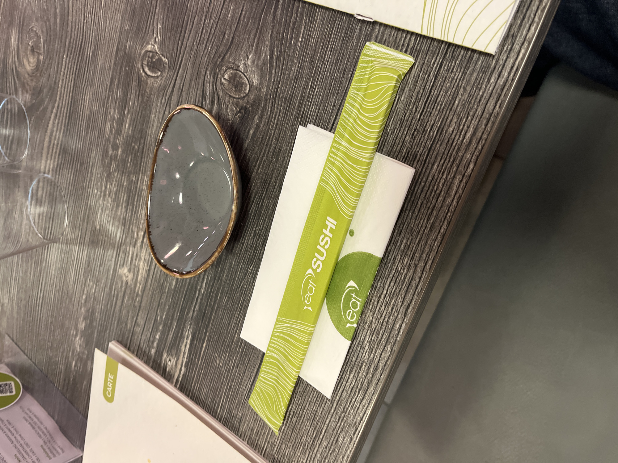The image shows a tabletop setting with a gray, wooden-patterned surface. A small, round, gray bowl with a gold rim is positioned near the top left. Next to it, slightly angled towards the right edge, there's a white napkin with a green paper wrapper on top, printed with "eat SUSHI" in a vertical arrangement. A menu card is visible in the lower left corner, with the word "CARTE" printed on its edge. Further to the left are parts of a wine glass. A section of a table or countertop appears in the lower right corner.
This scene takes place at the eat sushi restaurant, located in Andrézieux-Bouthéon, France, specifically at 25 Av. des Monts du Soir.