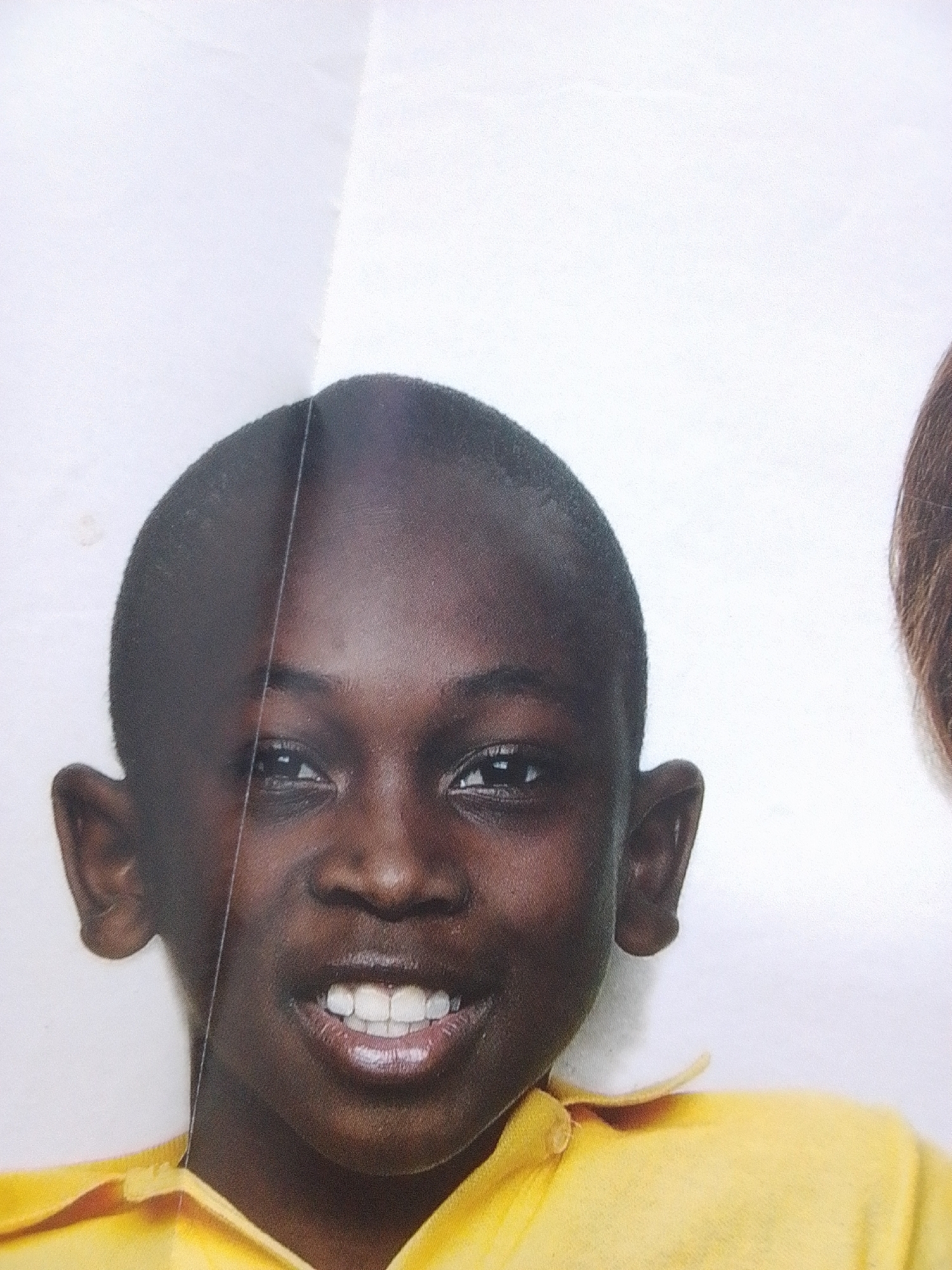 This media file is a close-up, head-and-shoulders photograph of a young Black boy. He is wearing a bright yellow collared shirt with a button at the neck. The background is a plain white, suggesting a studio setting or a well-lit indoor environment. The boy has dark skin, short-cropped hair, and prominent ears. He is smiling widely, showing a full set of white teeth. His eyes are dark and appear to be looking directly at the camera. A thin vertical line, possibly a crease or a fold in the background material, runs down the left side of his face, slightly obscuring the left side of his forehead and eye. To the right of the boy, a portion of another person's head and hair, appearing lighter brown, is visible, though out of focus. There is no visible text in the image. The lighting is bright and even, casting minimal shadows, which suggests it might be daytime or professionally lit. The overall impression is of a cheerful and direct portrait of a young individual. The location context provided is La Tagua, Venezuela.