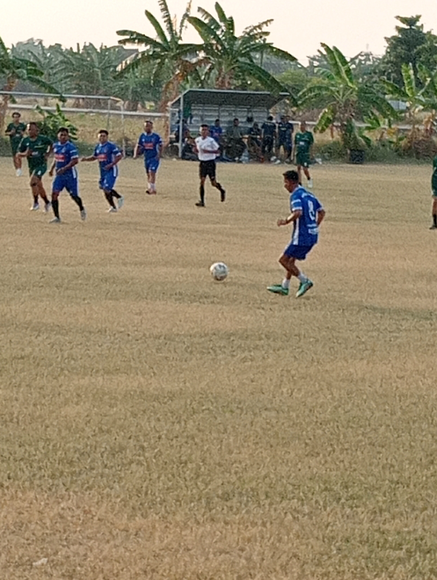 The image depicts a soccer game being played on a field in Kenep, Indonesia. Several players in blue and green uniforms are visible on the field. In the foreground, a player in a blue uniform is kicking a soccer ball. In the background, there are trees and a small building, possibly a shelter or changing room. The field appears to be made of dry grass. The sky is overcast, suggesting it might be late afternoon or early morning.